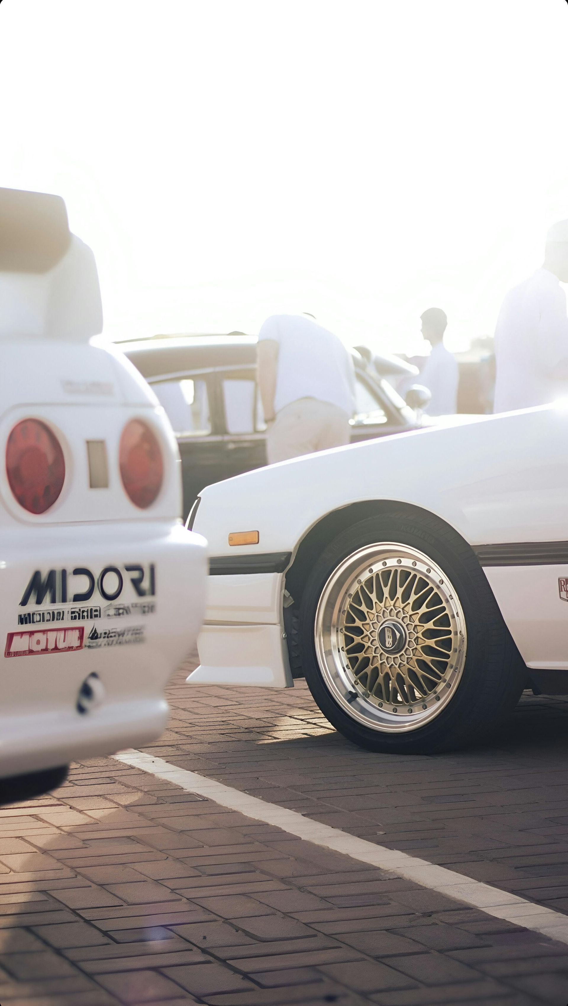 Close-up of white sports cars parked on a brick lot, featuring a Midori sticker on the rear bumper and gold-rimmed wheels.