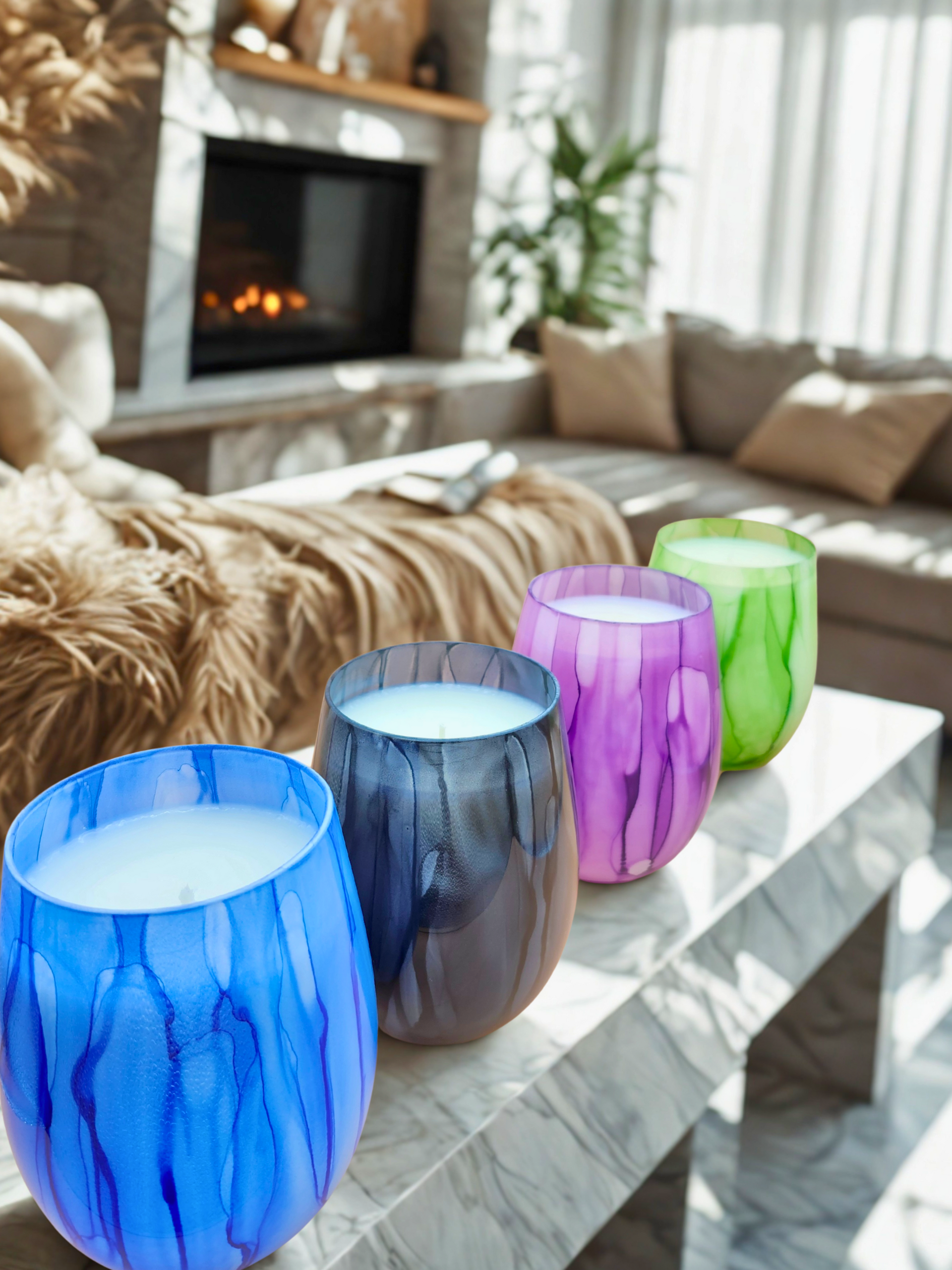 Four colorful glass candles on a white marble coffee table; blurred living room with fireplace in background.