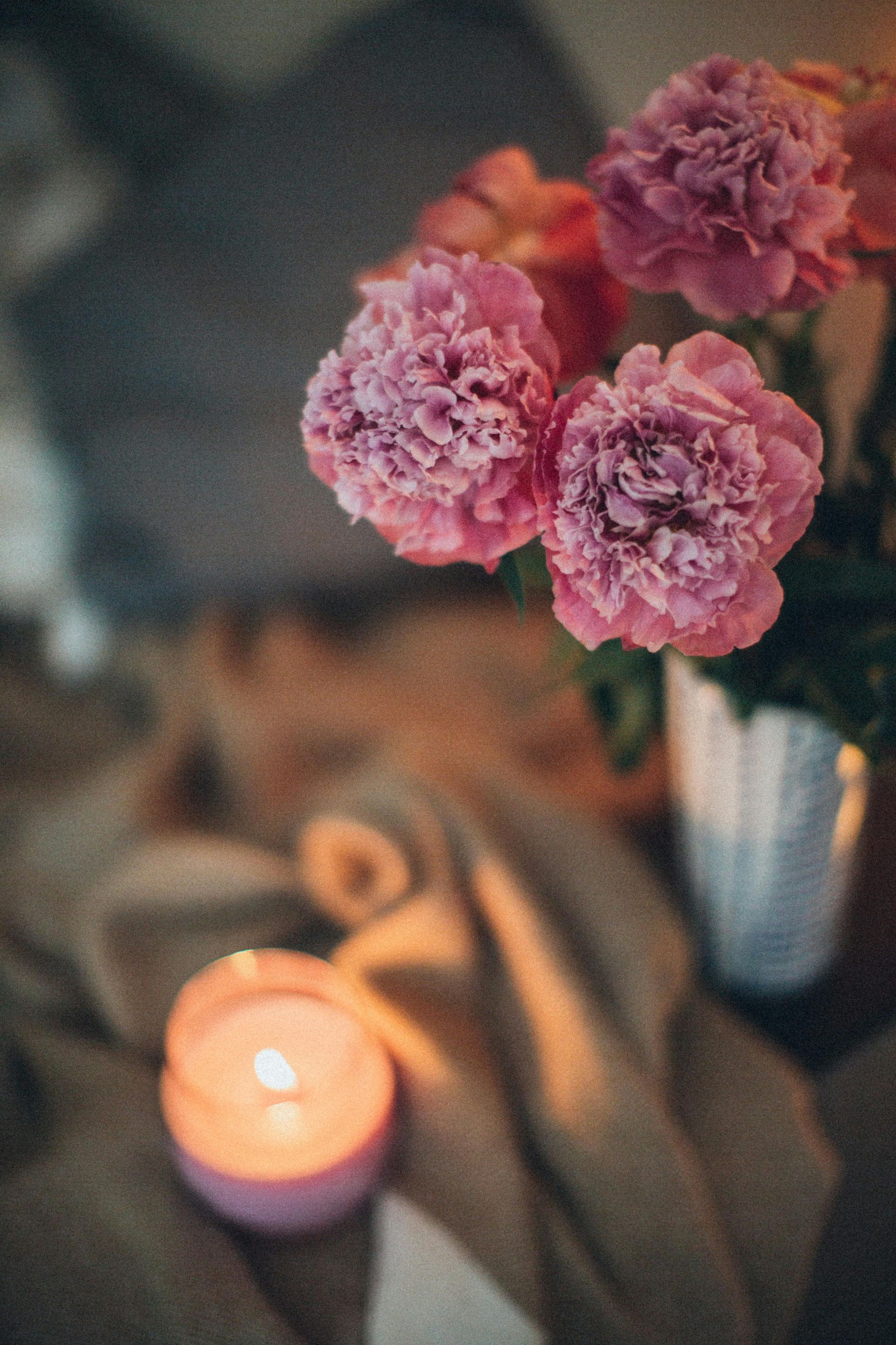 Pink peonies and a lit candle on a draped surface create a cozy, warm ambiance.