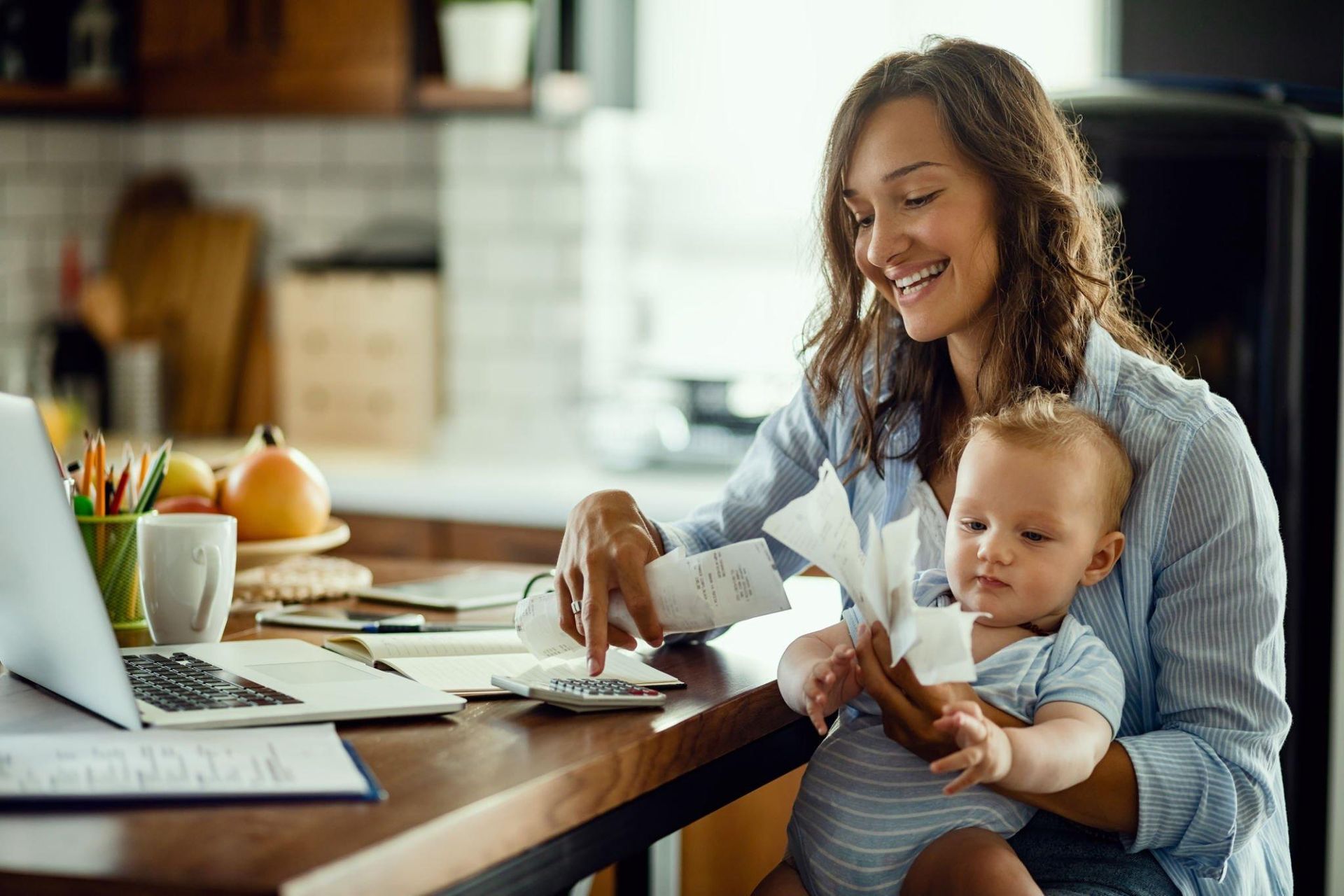 A woman is holding a baby while sitting at a table with a laptop.