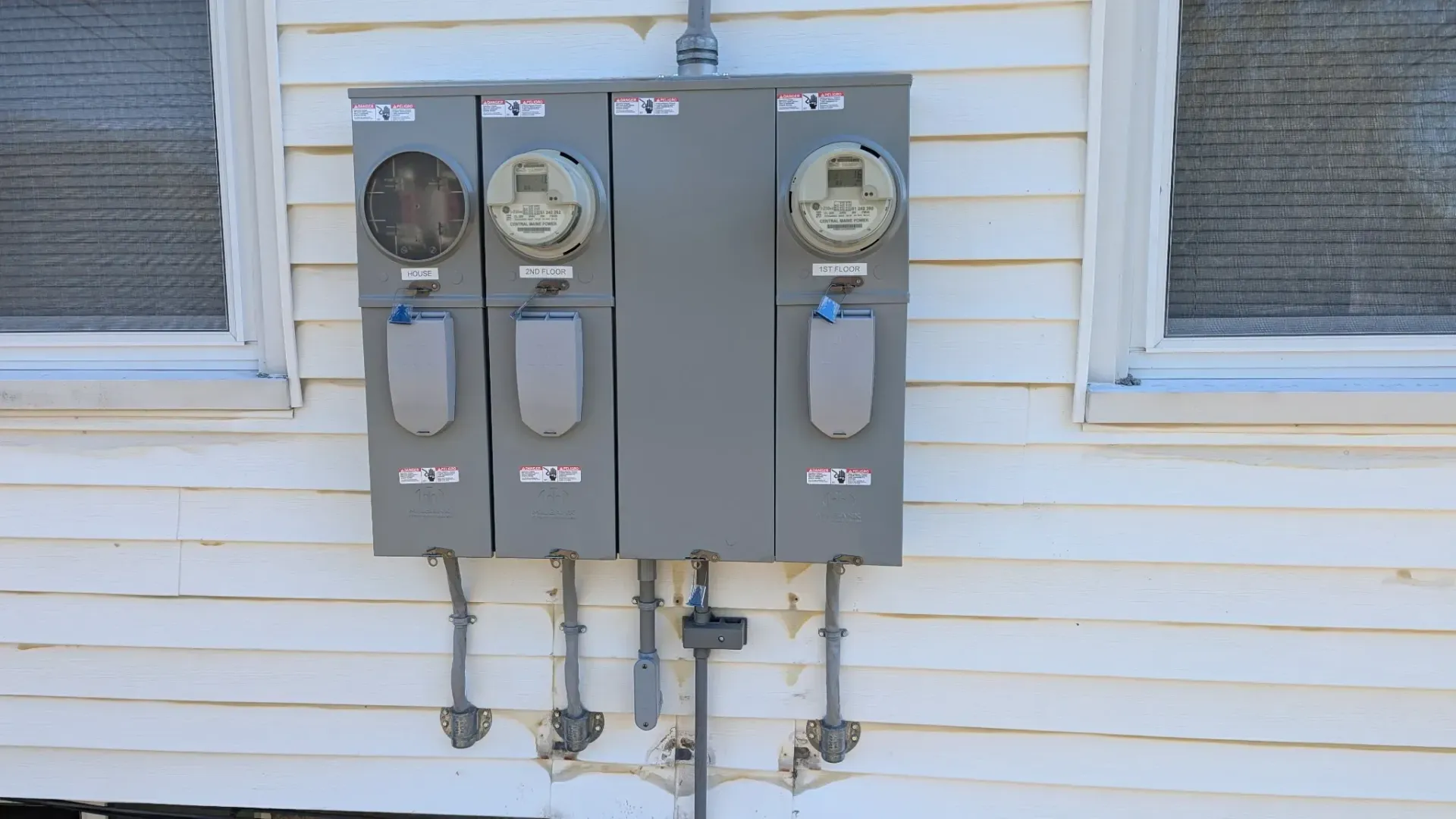 Three electric meters mounted on a white, clapboard house exterior. Gray metal boxes and conduits.