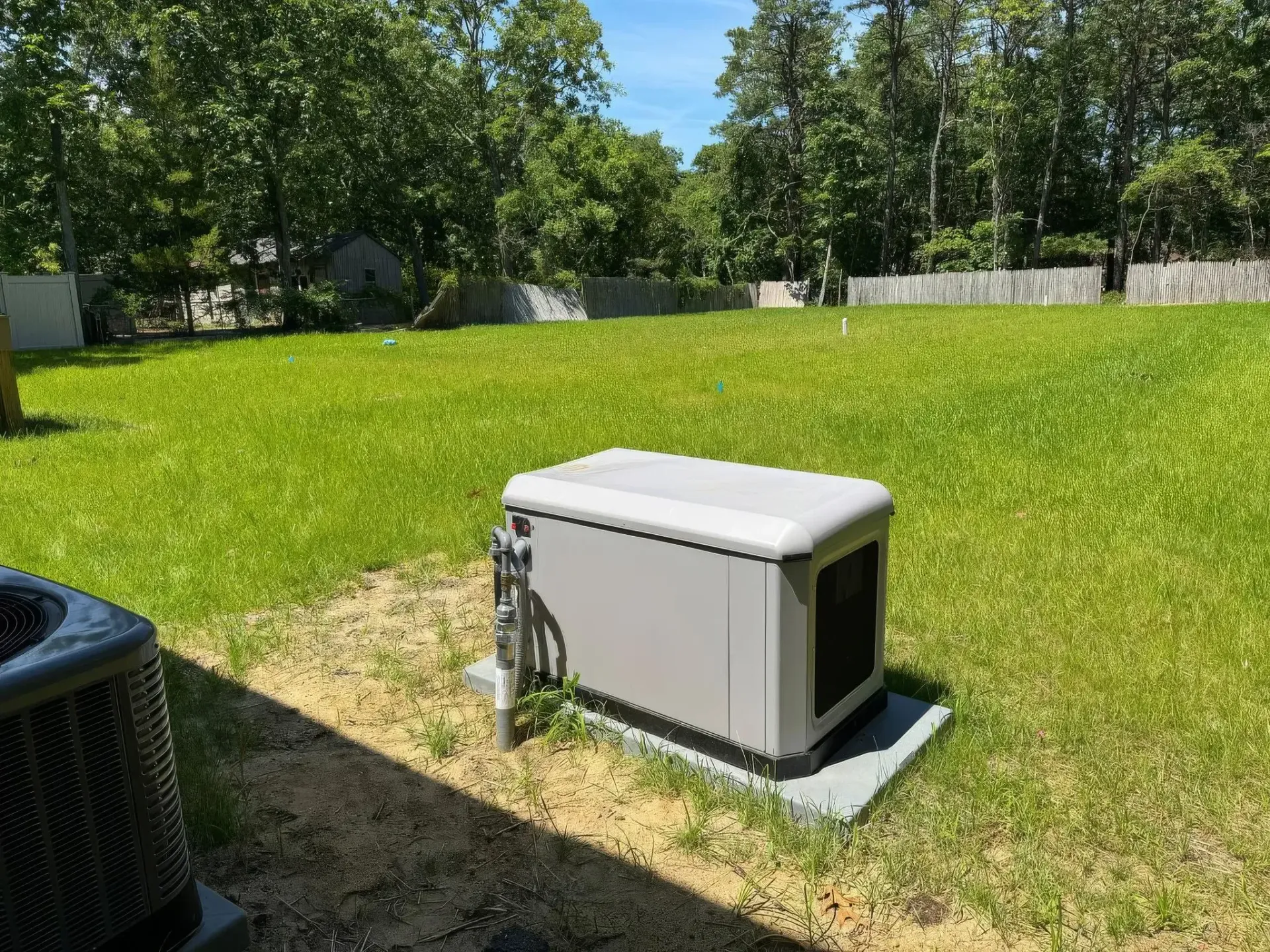 Gray generator on concrete pad in grassy yard, surrounded by trees and a fence.