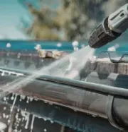 Pressure washing a roof gutter with a spray nozzle, water splashing over dark shingles