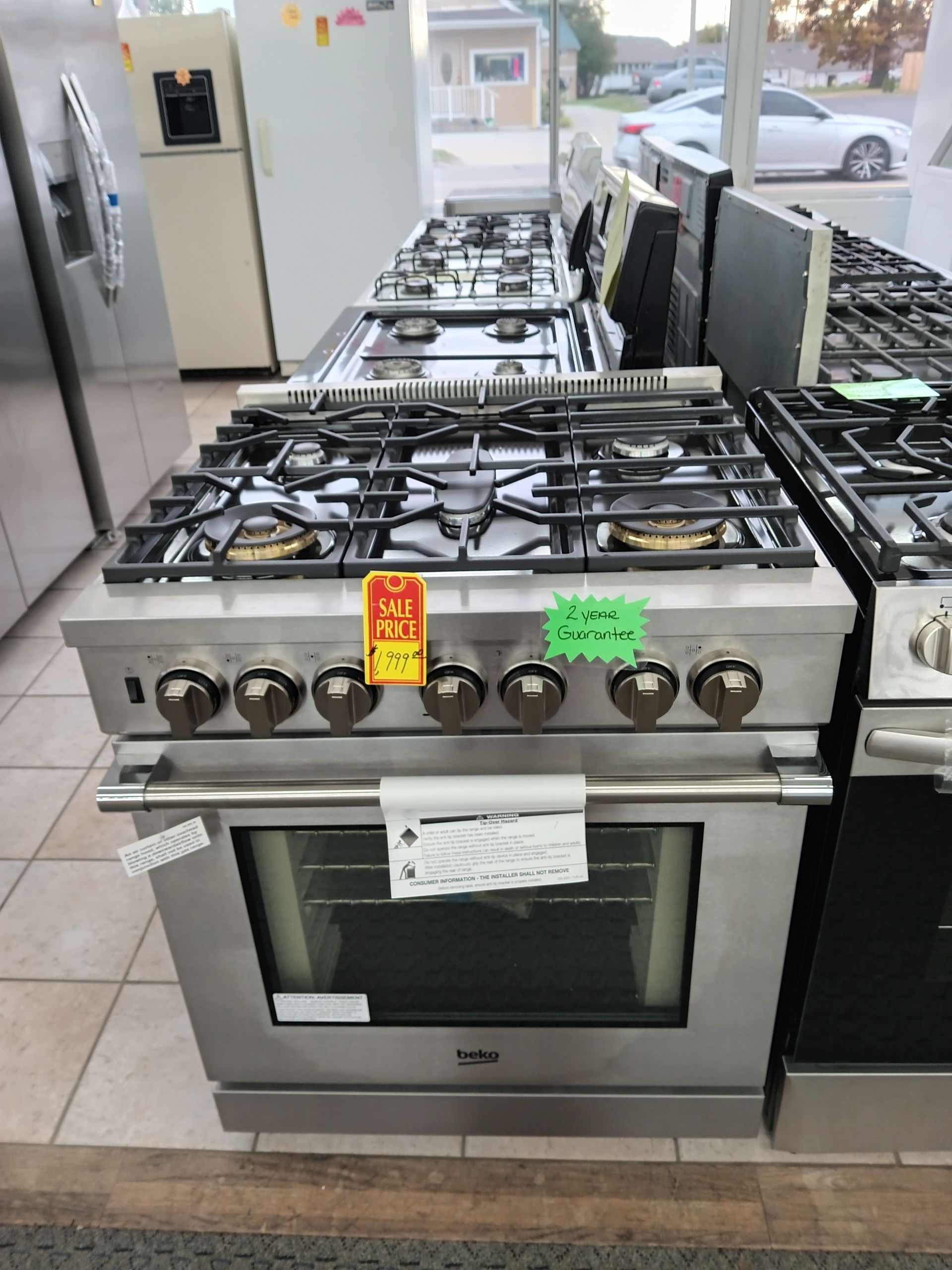 Stainless steel gas range in a showroom, with other appliances in the background.