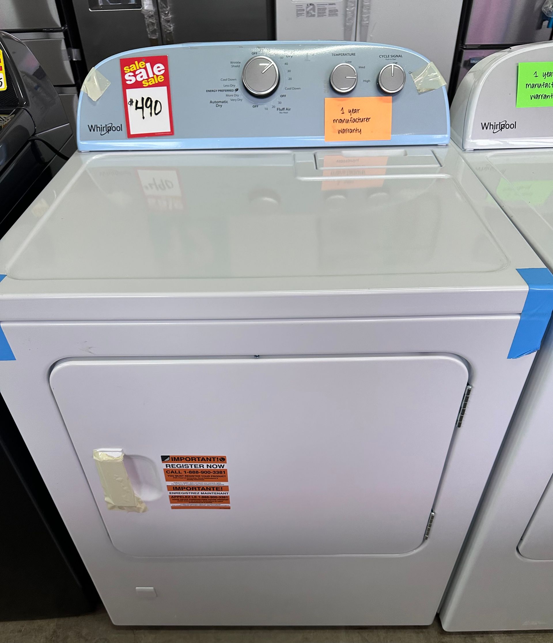 A white clothes dryer with a light blue top control panel, displayed in a retail store with sale tags attached.