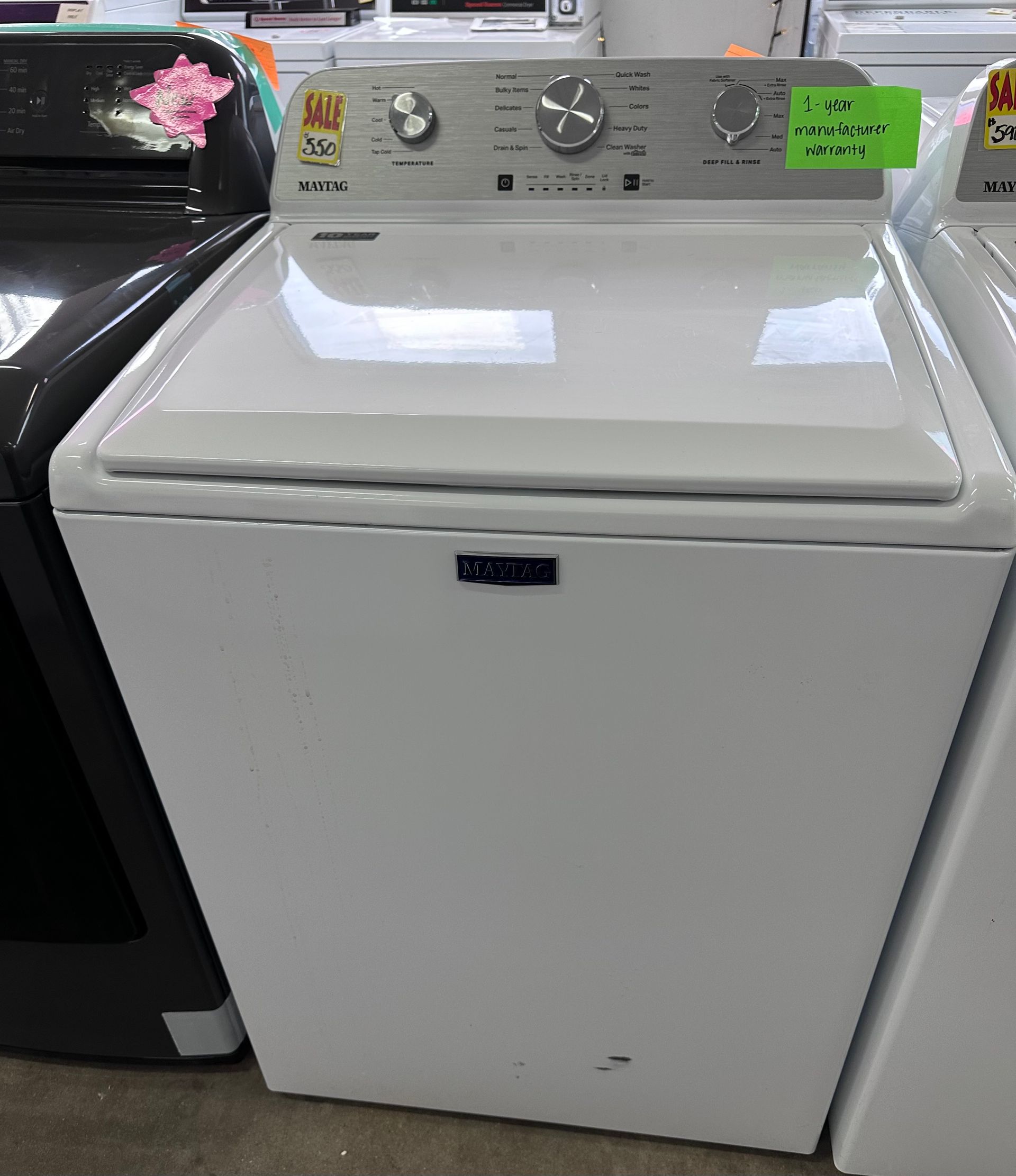 A white top-loading Maytag washing machine with a silver control panel, displayed in a retail showroom.