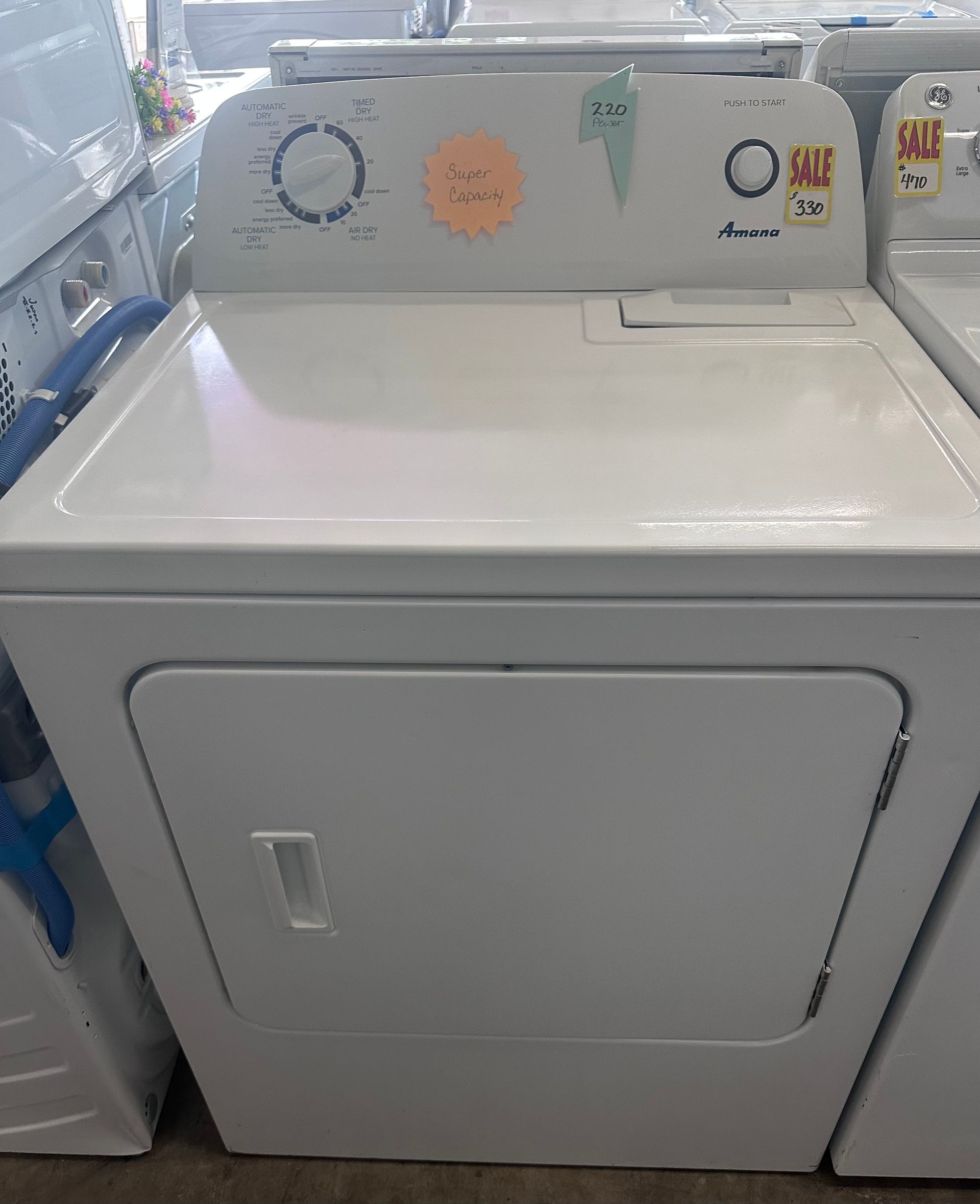 White Amana dryer with top controls and a front door, in a store setting.