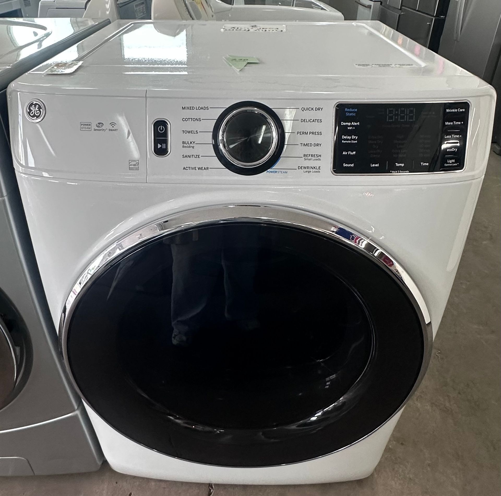 A slate-gray Maytag front-load clothes dryer with a black glass door, displayed on a retail showroom floor.