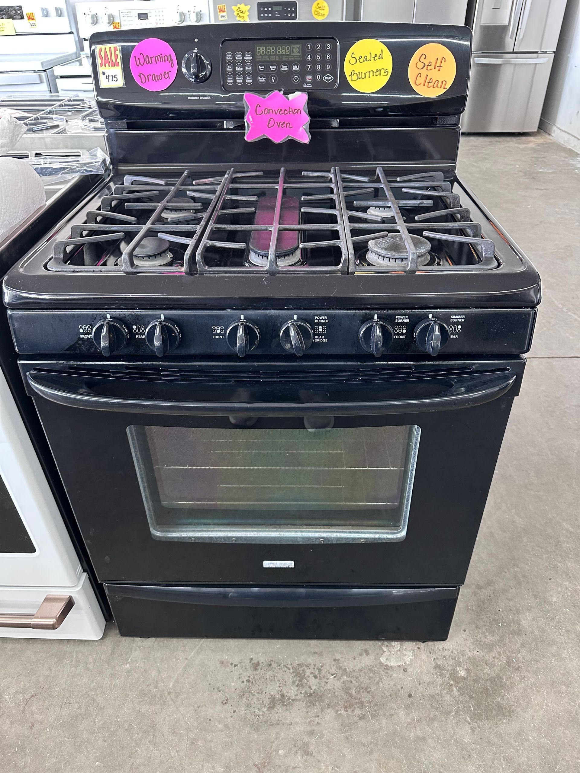 A black gas range with a stovetop and oven, featuring several colorful price stickers on the back control panel.