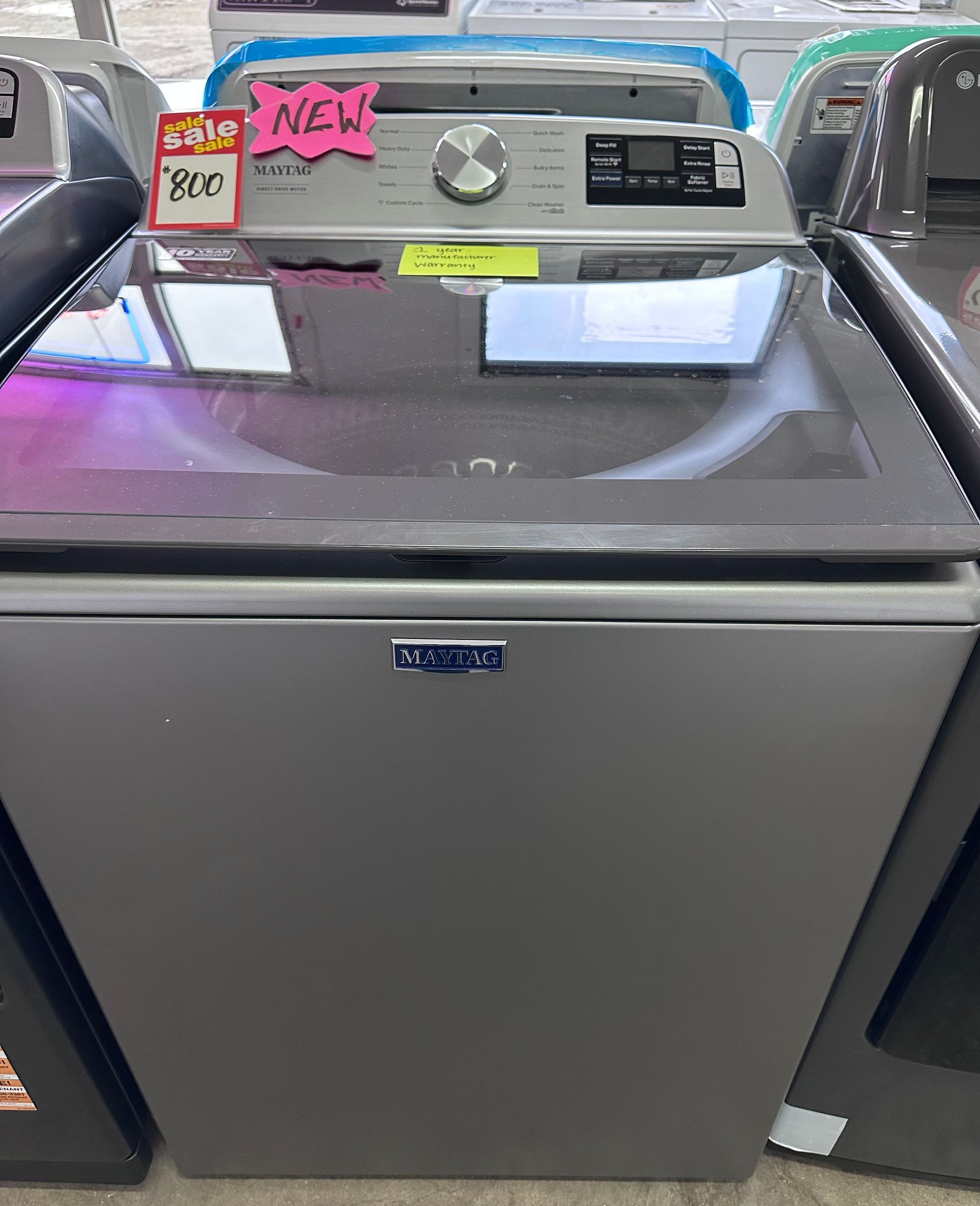 A metallic grey Maytag top-load washing machine with a glass lid and digital control panel on display in a store.