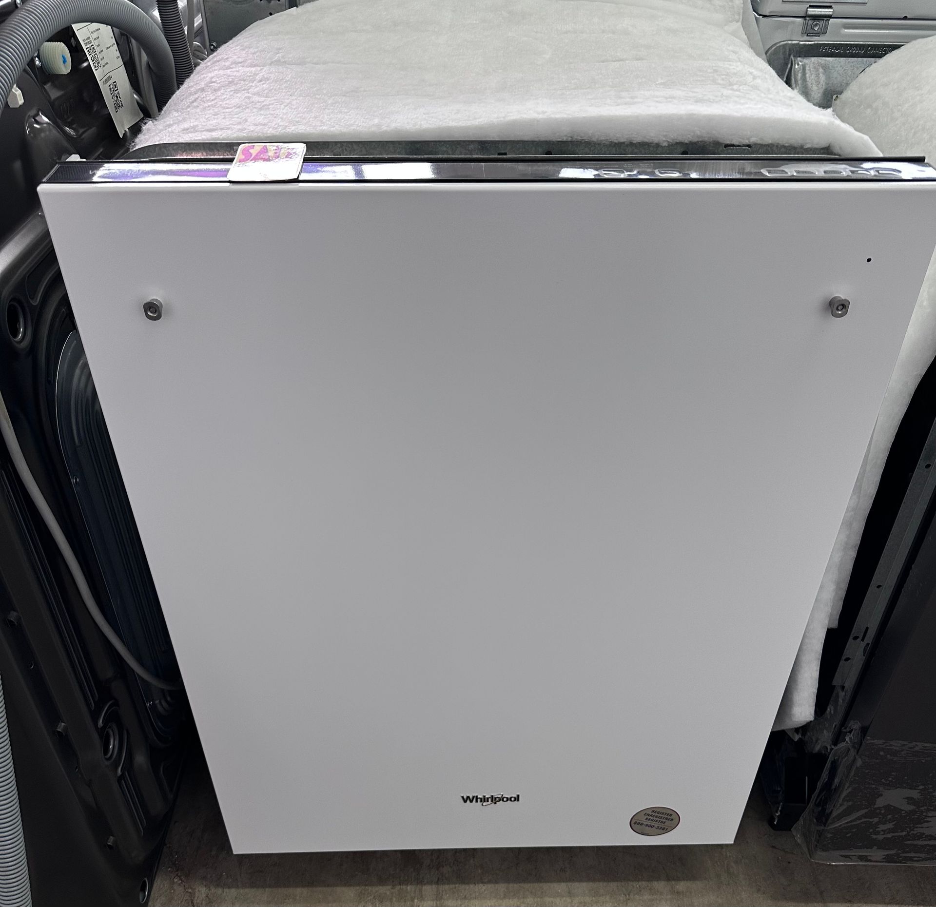 A white front-loading Whirlpool dishwasher with a protective plastic film on top, situated in a warehouse or store.