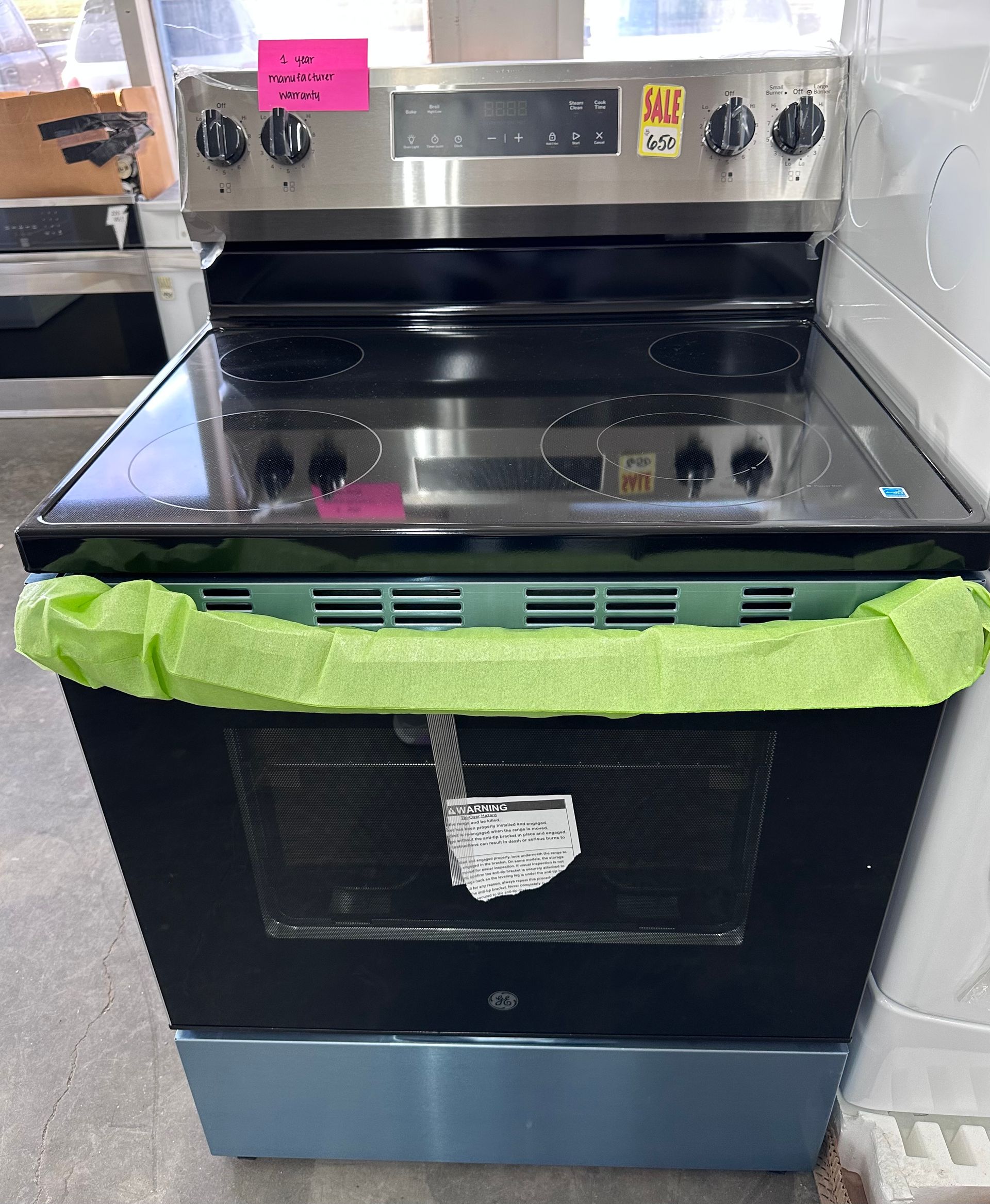 A stainless steel electric range with a black glass cooktop and oven door, wrapped in green protective foam packaging.
