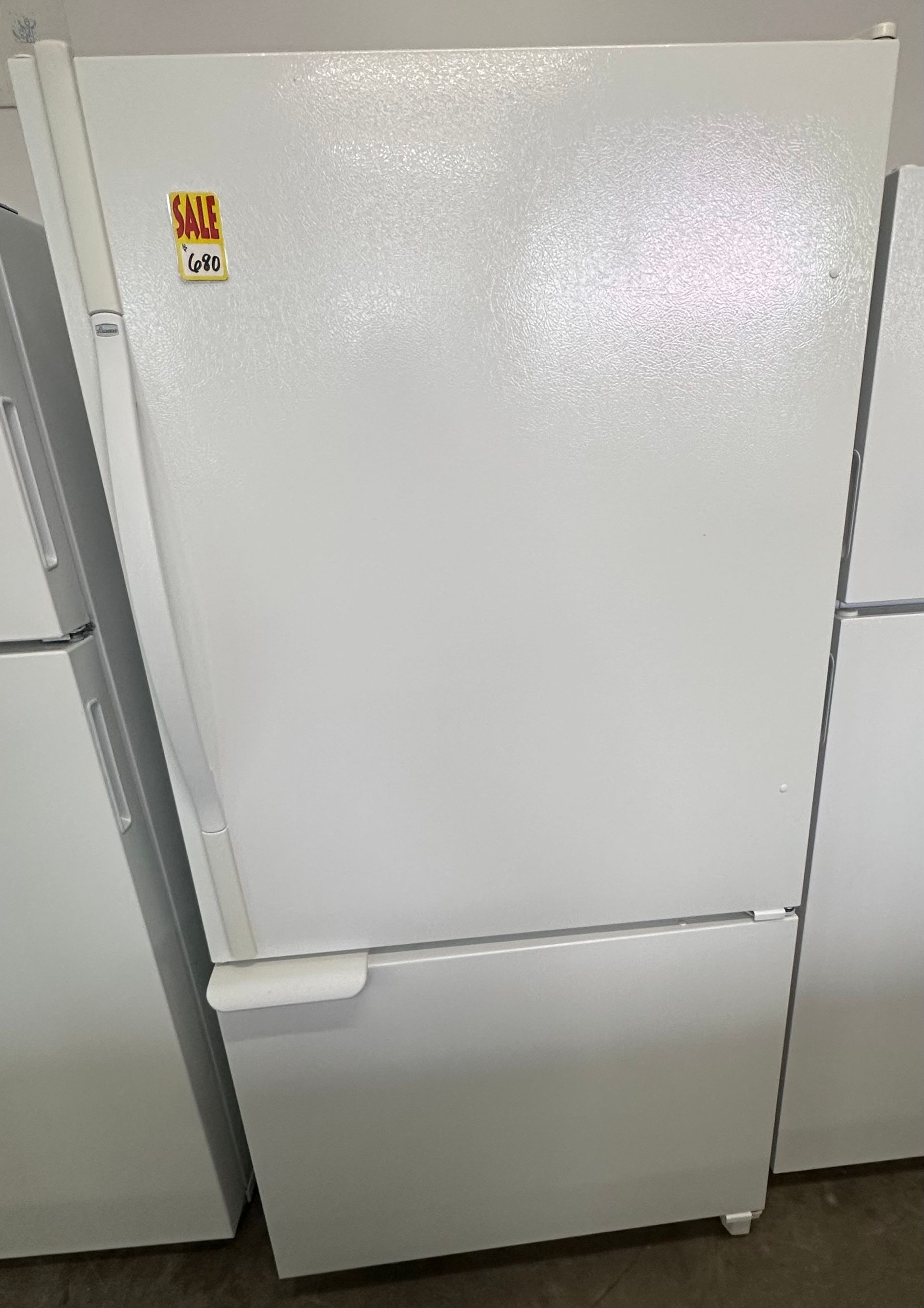 A tall, dark-colored refrigerator with stickers on the door, set against a white wall and a dark floor.