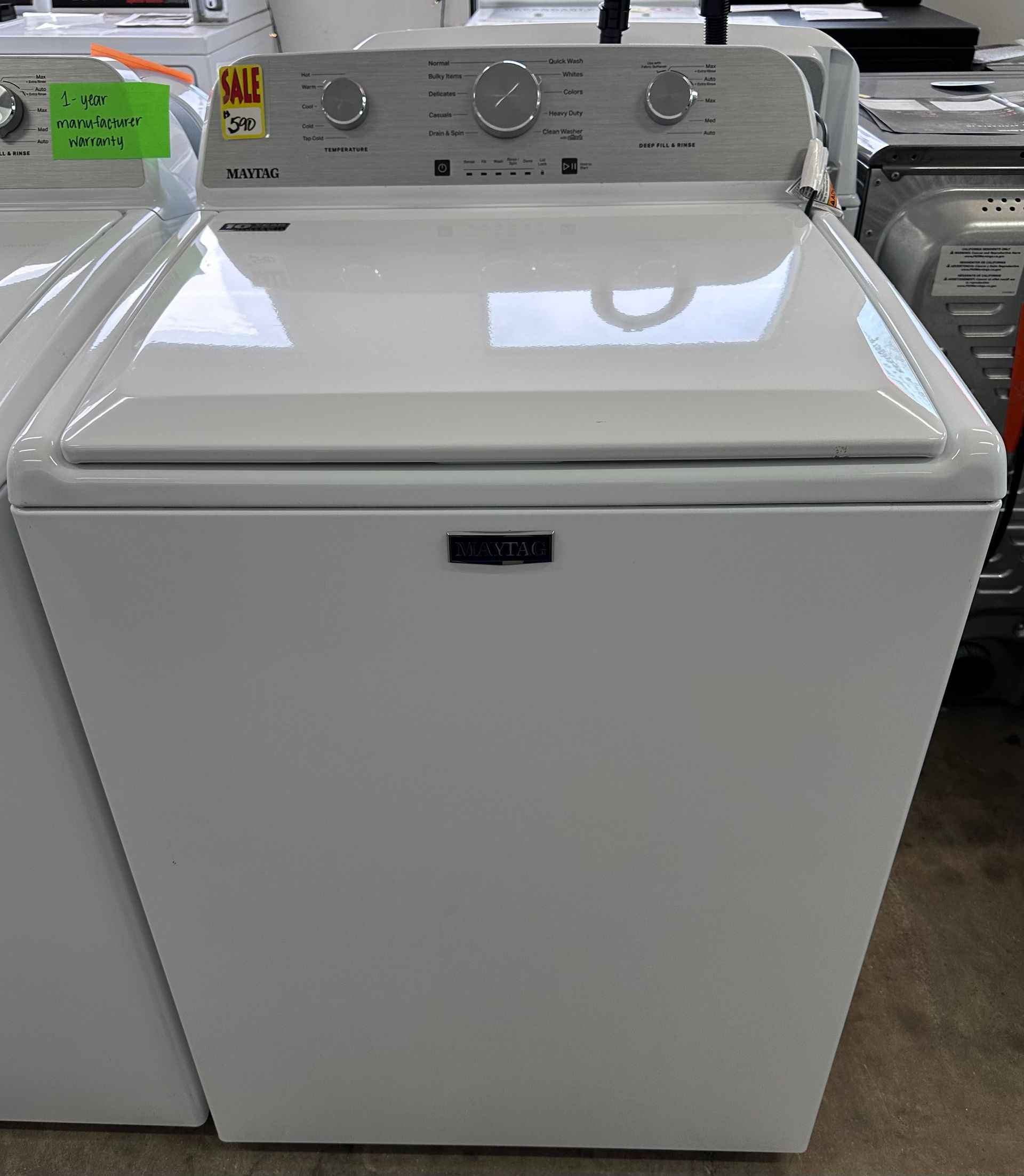 A white Maytag top-loading washing machine with a control panel on the back, situated in a store retail setting.