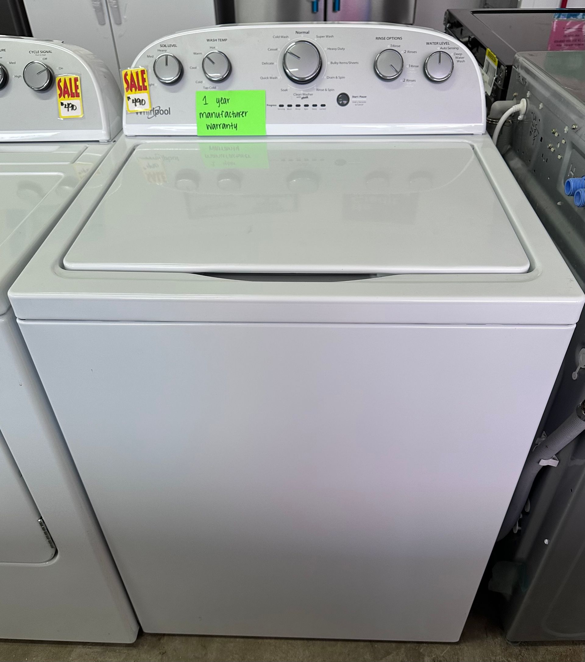 A white top-loading washing machine with a control panel featuring five knobs, displayed in a retail showroom.