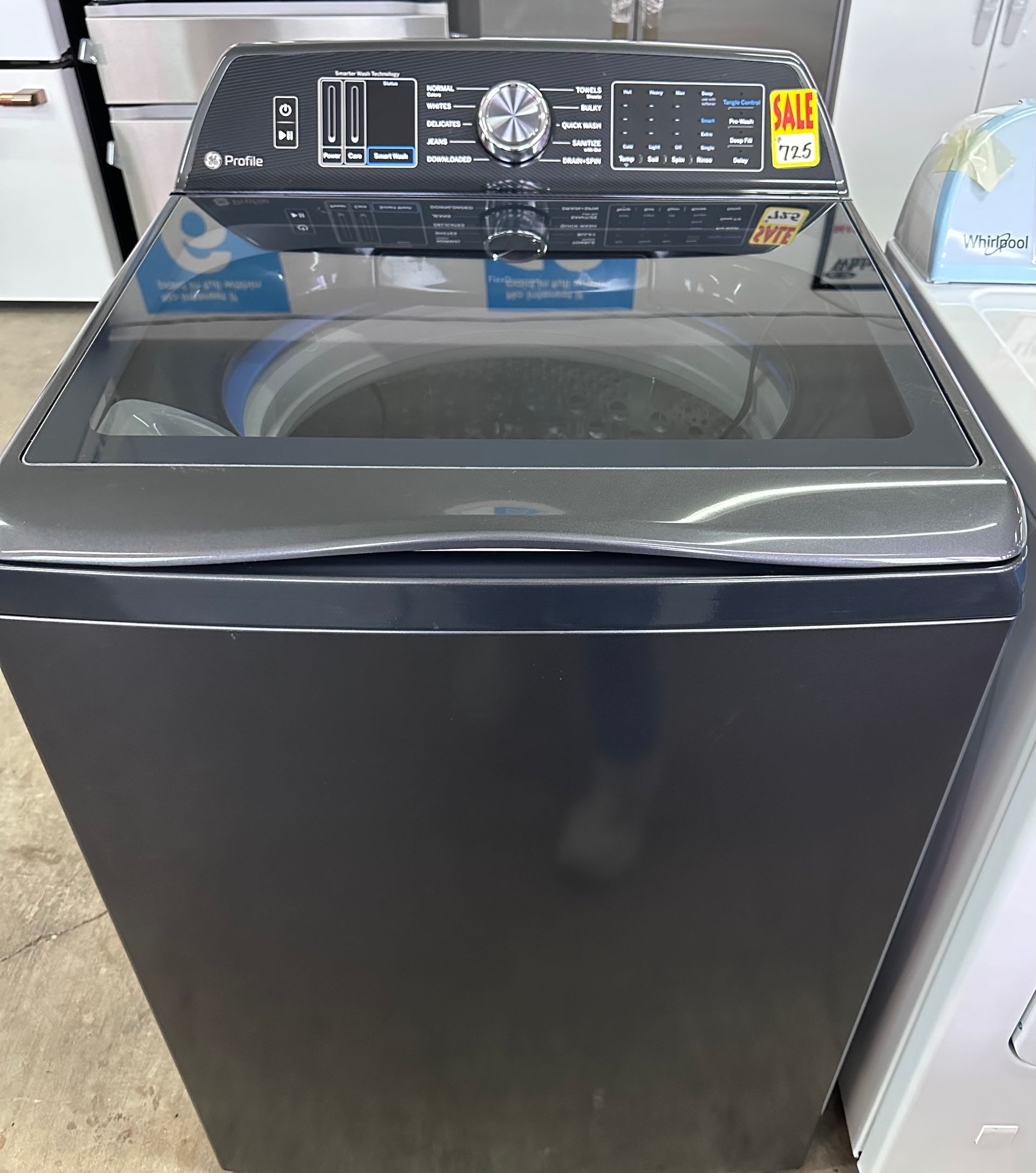 A dark grey top-loading washing machine with a black glass lid and a control panel with a central dial.