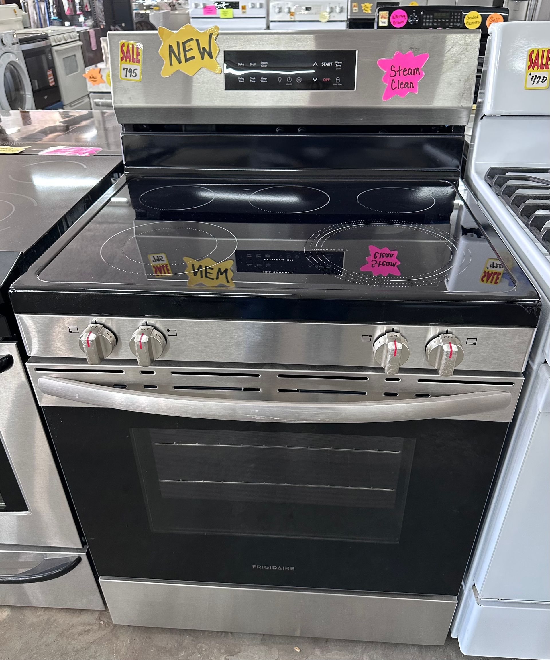 A stainless steel electric range with a black glass cooktop and oven door, displayed in a retail showroom.