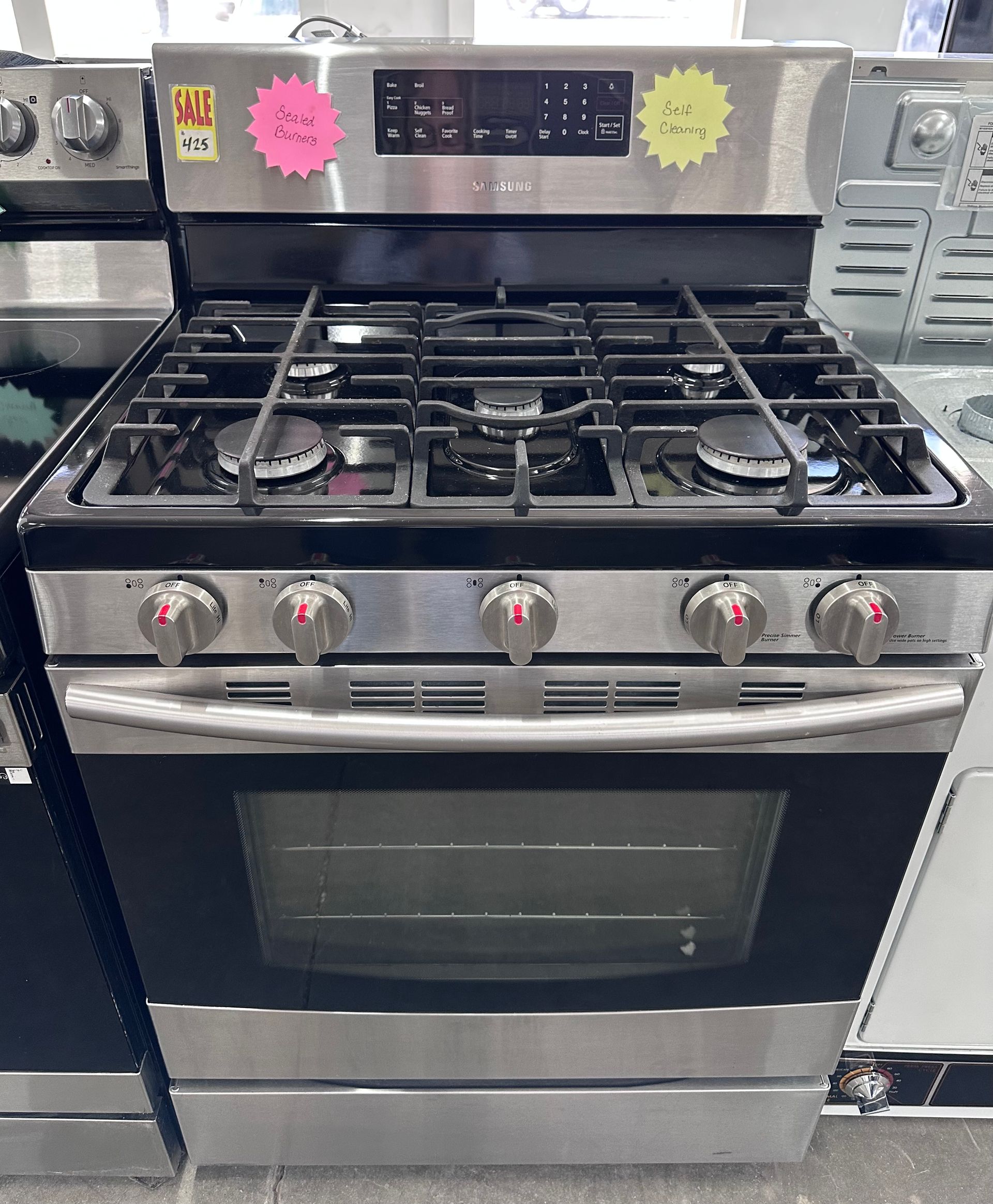 A stainless steel, free-standing gas range with five burners, a front control panel, and an oven door in a store setting.