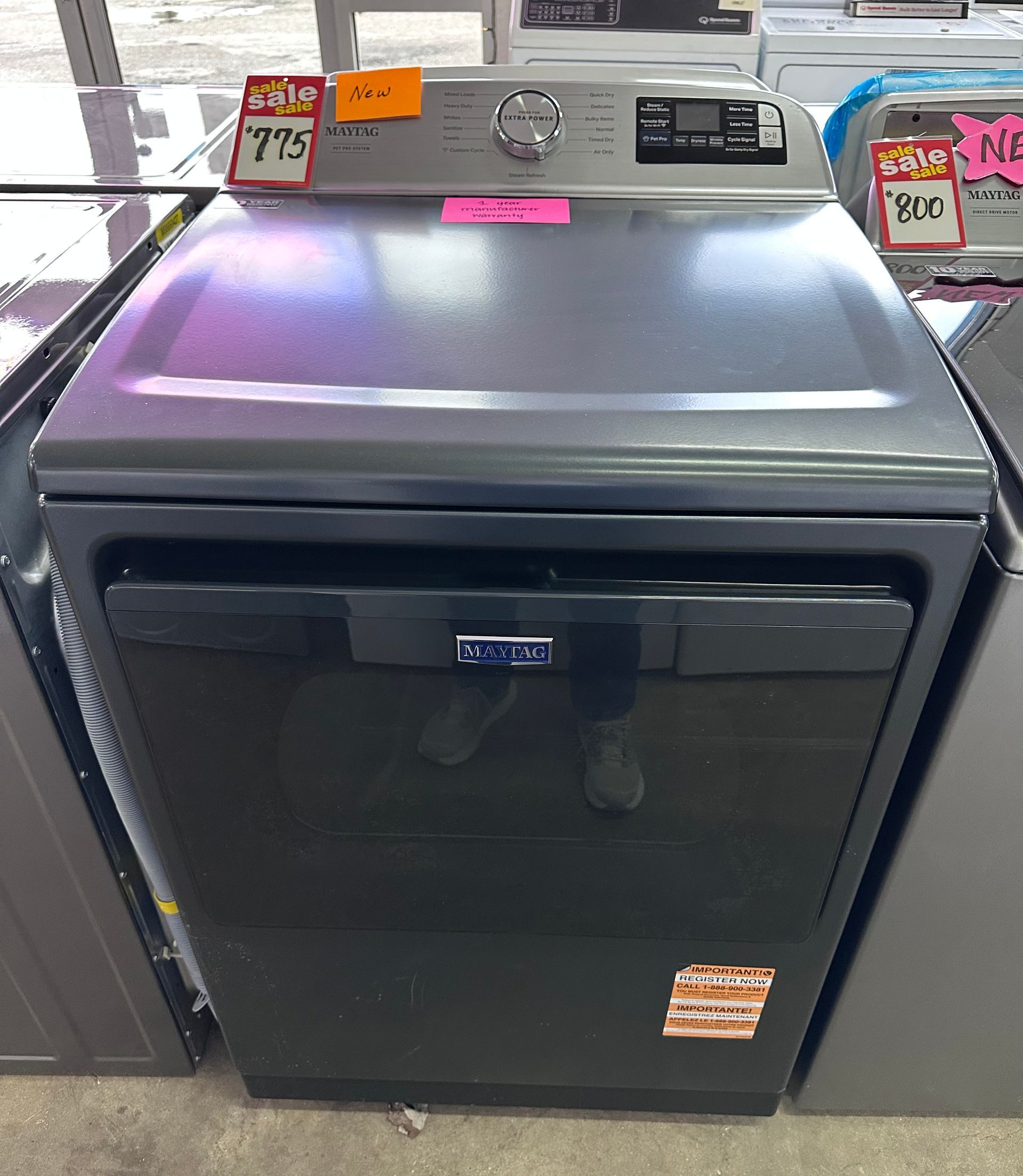 A slate-gray Maytag front-load clothes dryer with a black glass door, displayed on a retail showroom floor.