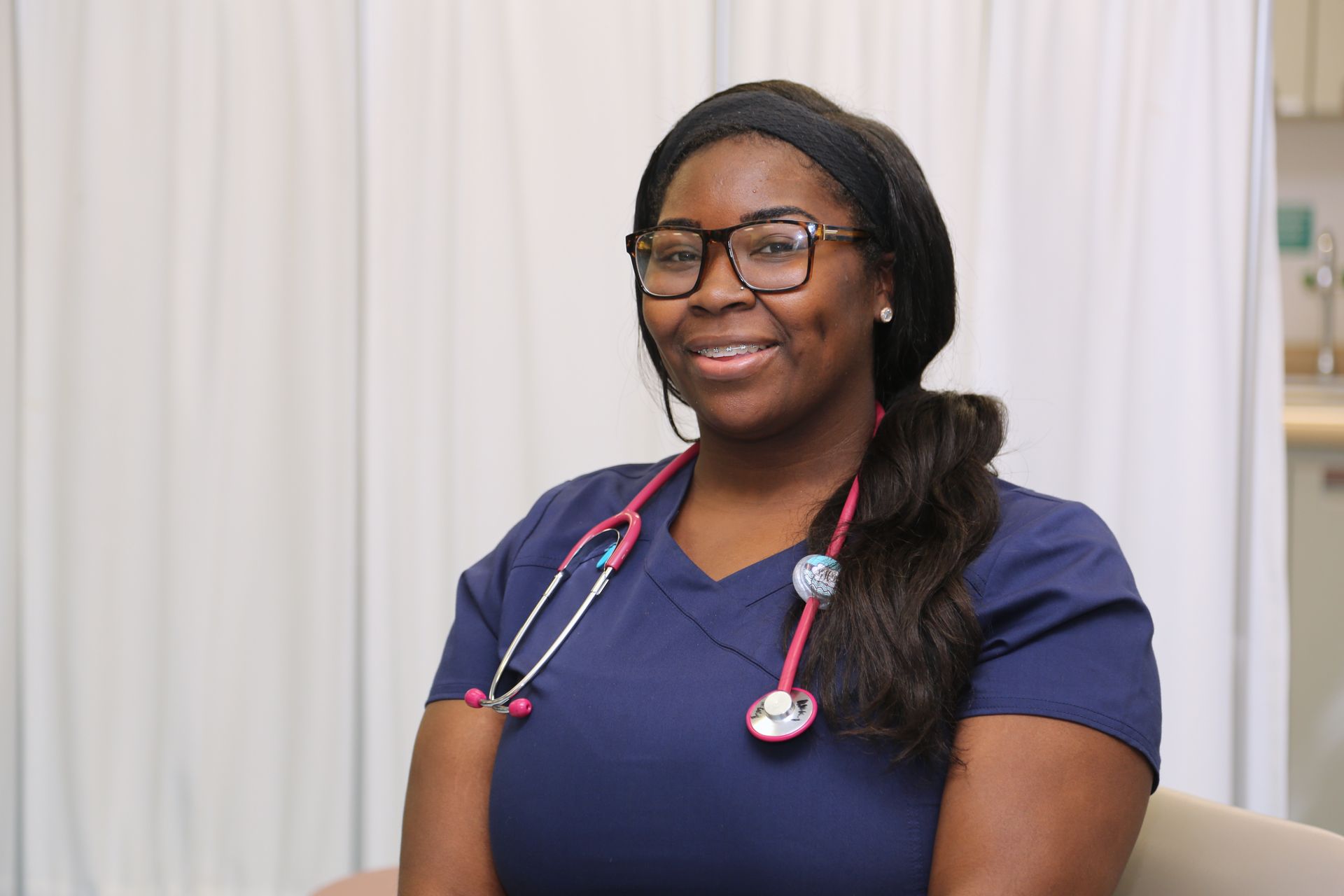 Nurse in blue scrubs with pink stethoscope, smiling, wearing glasses, in a medical setting.