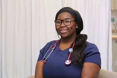 Nurse in blue scrubs with pink stethoscope, smiling, wearing glasses, in a medical setting.