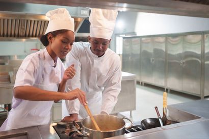 Two chefs in white uniforms stirring a pot on a stove in a commercial kitchen.