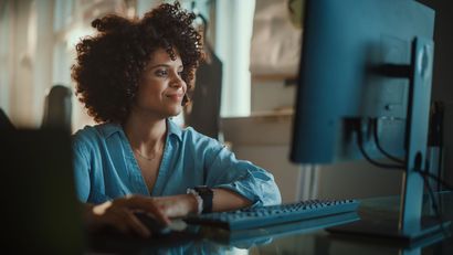 Woman with curly hair smiles while working on a computer in a sunlit office.