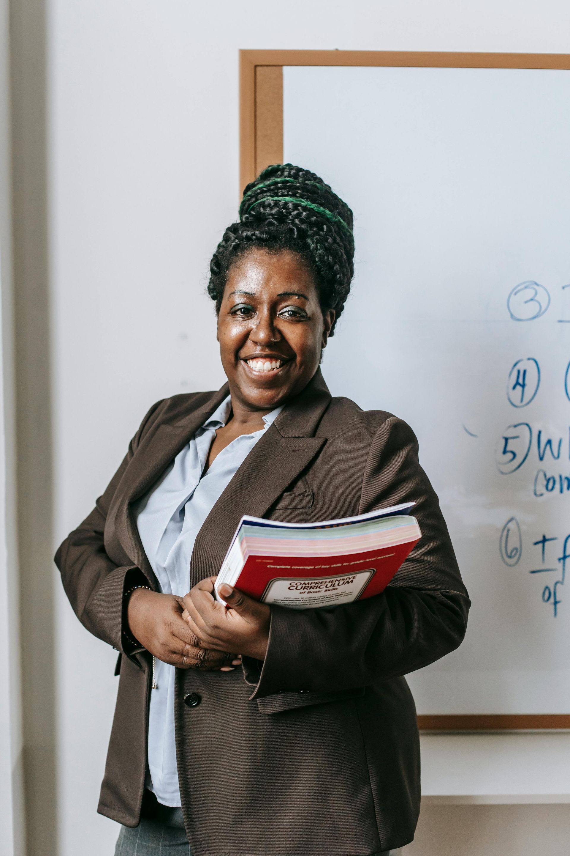 Woman in blazer holds a book, smiles in front of a whiteboard.