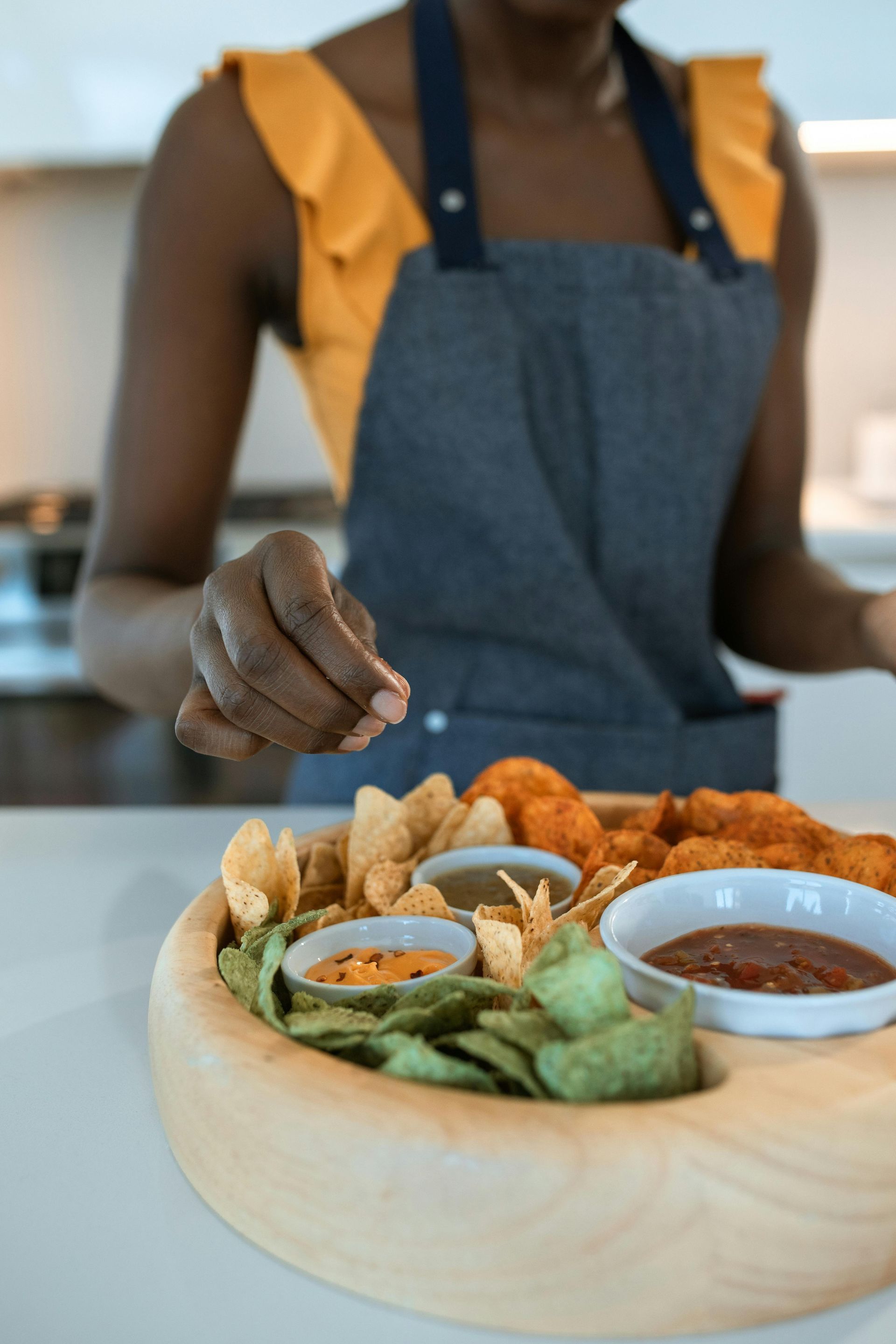 Woman seasoning food on a wooden platter with chips, dips, and chicken.