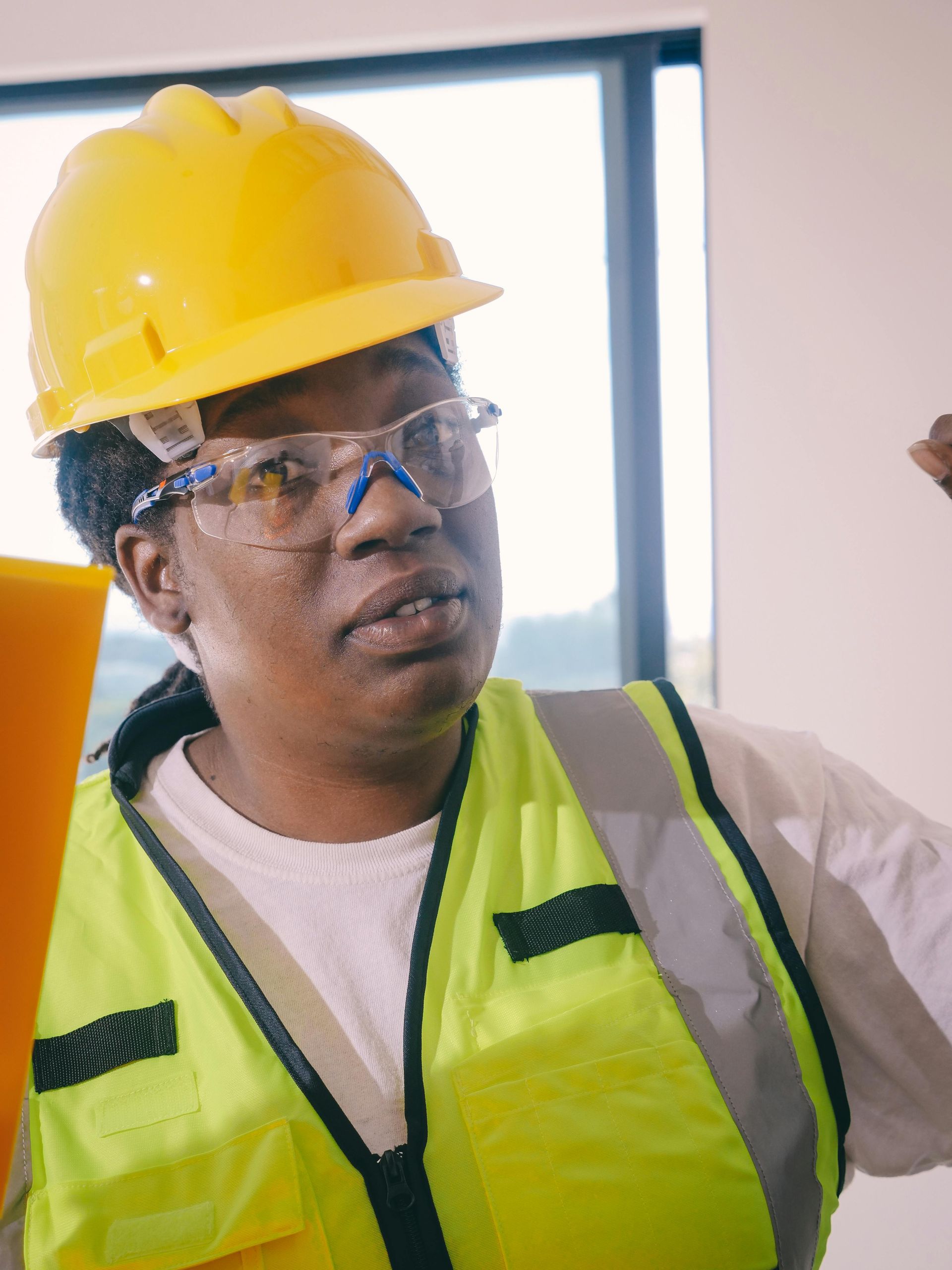 Woman in construction vest and hard hat smiles, leaning against a concrete wall.