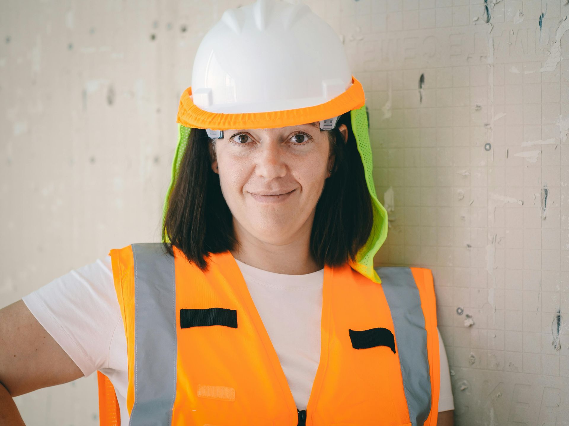 Woman in construction vest and hard hat smiles, leaning against a concrete wall.