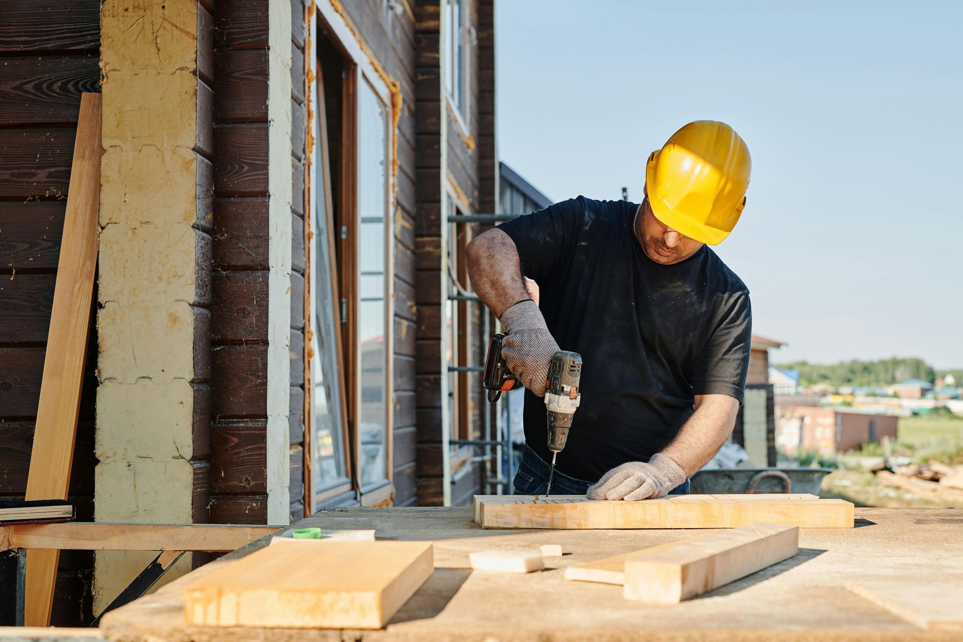 Construction worker in a yellow hardhat drilling into wood on a sunny day.