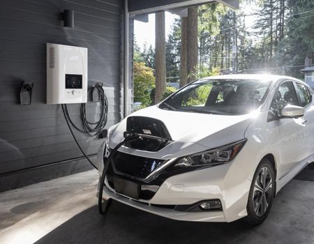 White electric car charging at a wall-mounted station in a garage with large windows.