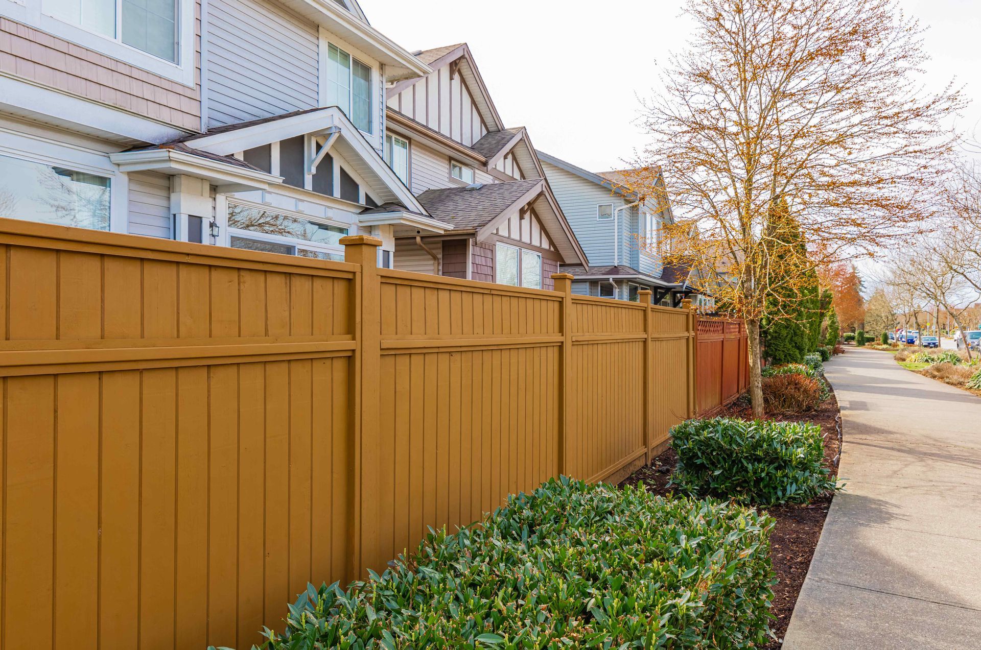 Brown wooden fence bordering a sidewalk and houses. Green bushes are in front.