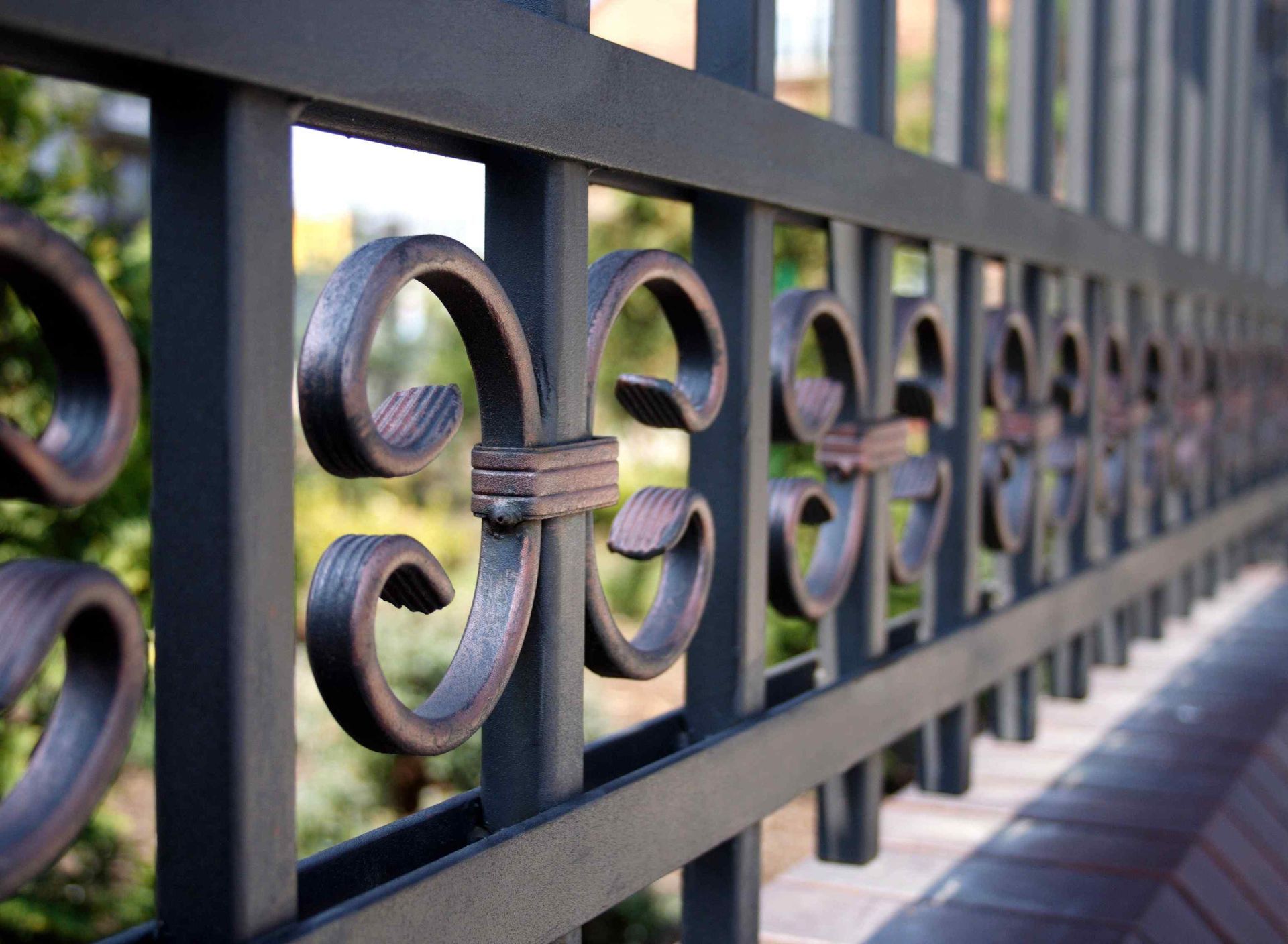 Close-up of a black wrought-iron fence with decorative scrollwork and a blurred background of greenery.