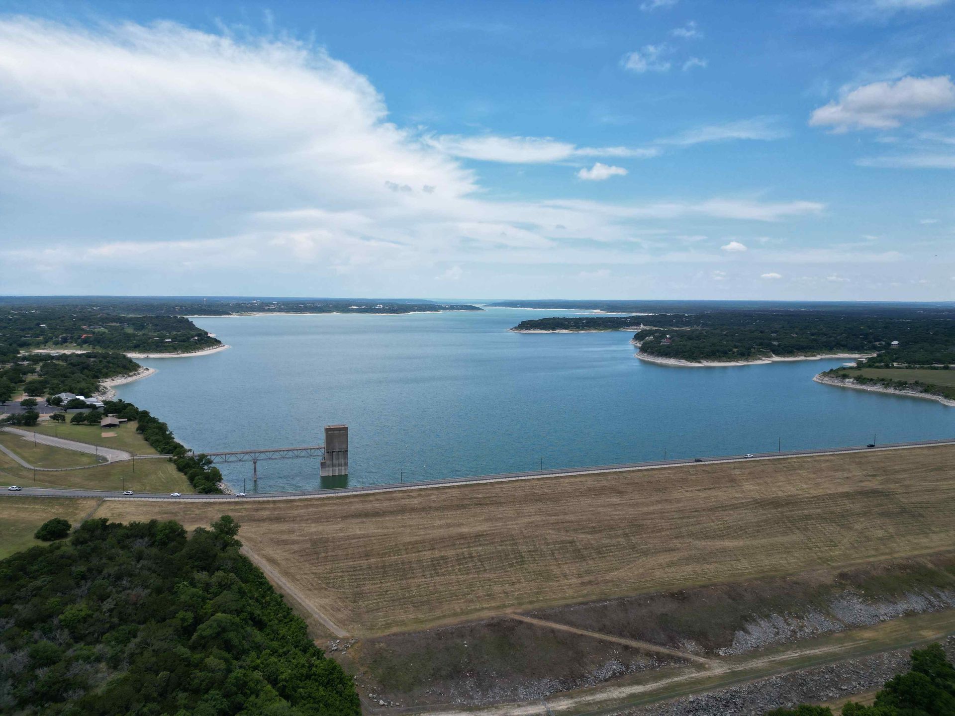 Blue lake behind a large dam, under a cloudy sky. Trees and buildings border the lake.
