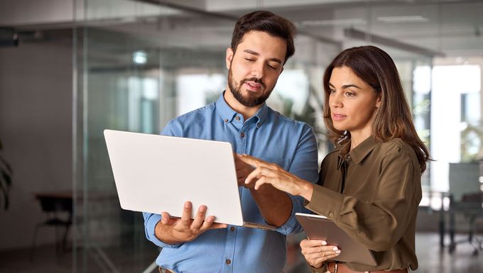 A man and woman in an office, looking at a laptop. The woman points to the screen while holding a tablet.