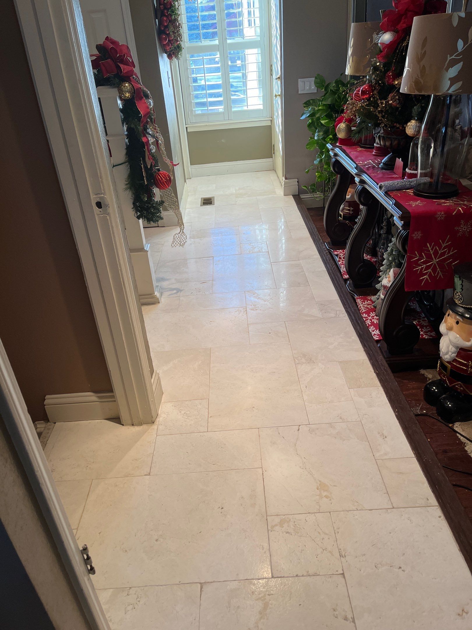 A hallway with light-colored tile flooring, leading toward a window and decorated with Christmas greenery and figures.