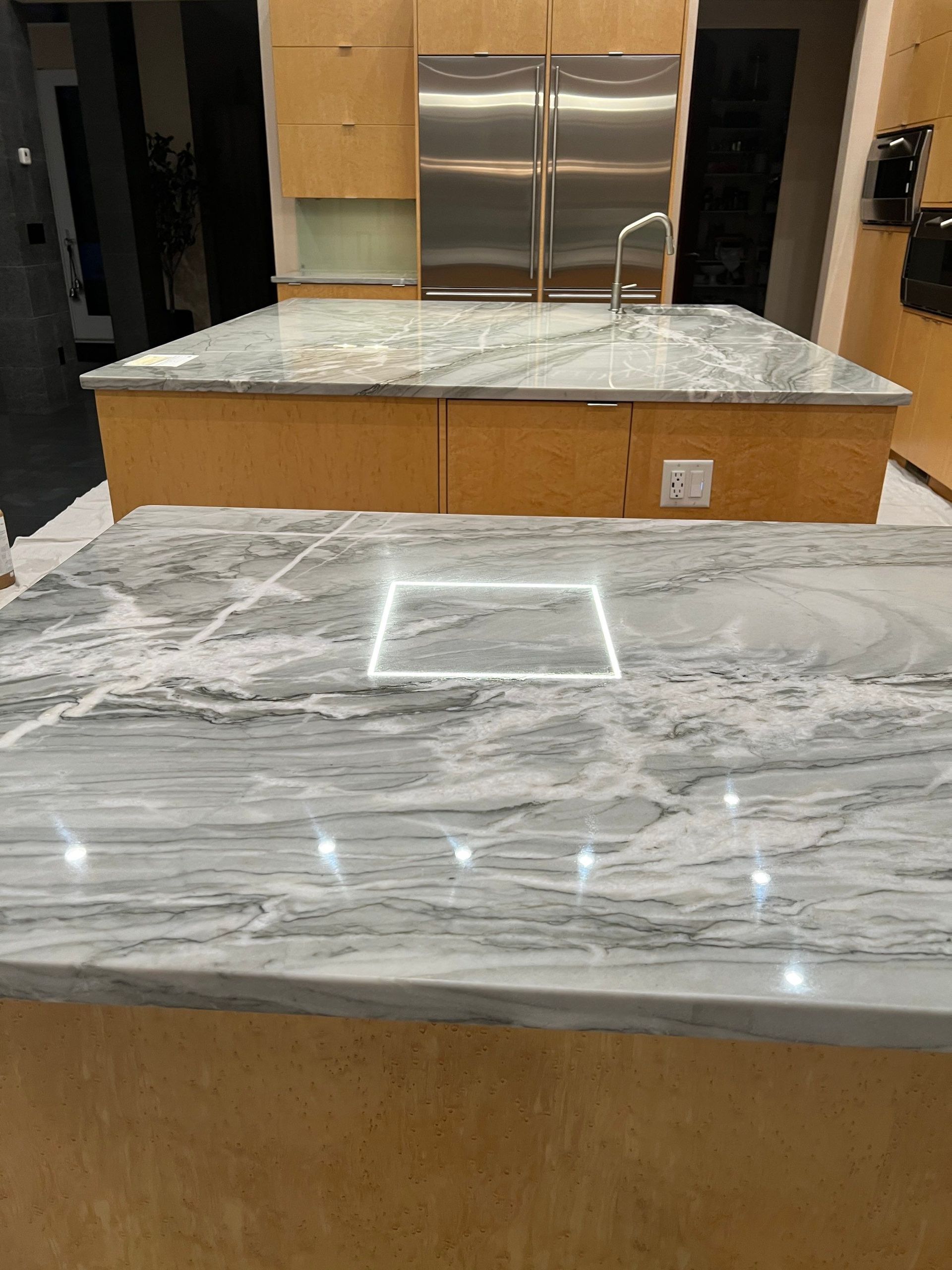 Marble countertops in a kitchen, with light fixtures on the island and stainless steel refrigerator.