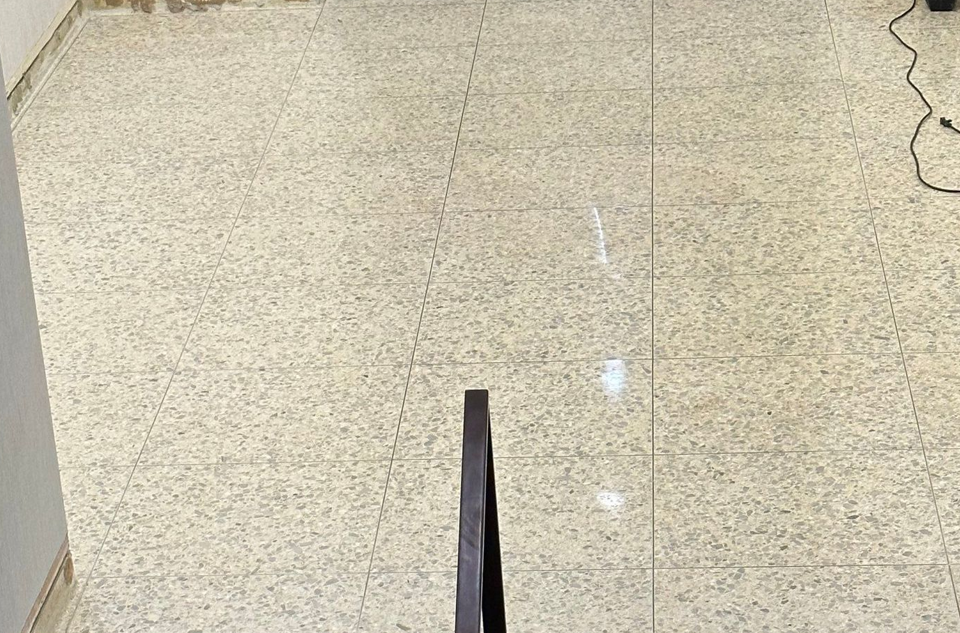 A high-angle view of a speckled light-colored floor, a vertical dark stair railing, and a faint line marking the floor.