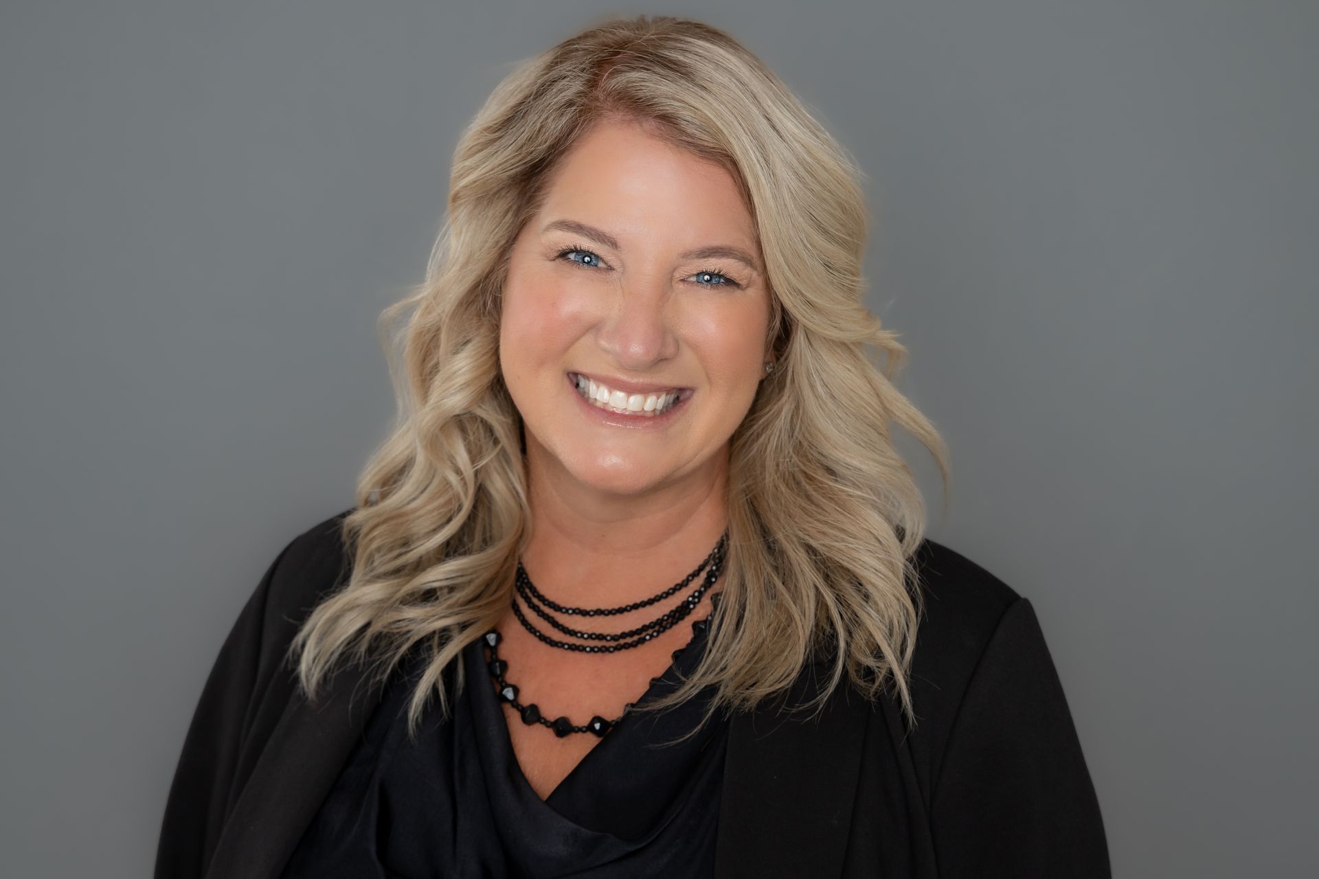 A woman wearing a black jacket and a necklace is smiling for the camera.