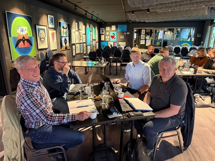 Gruppe menn ved et bord i et rom med kunst på veggene, noen ser på bærbare datamaskiner og papirer.