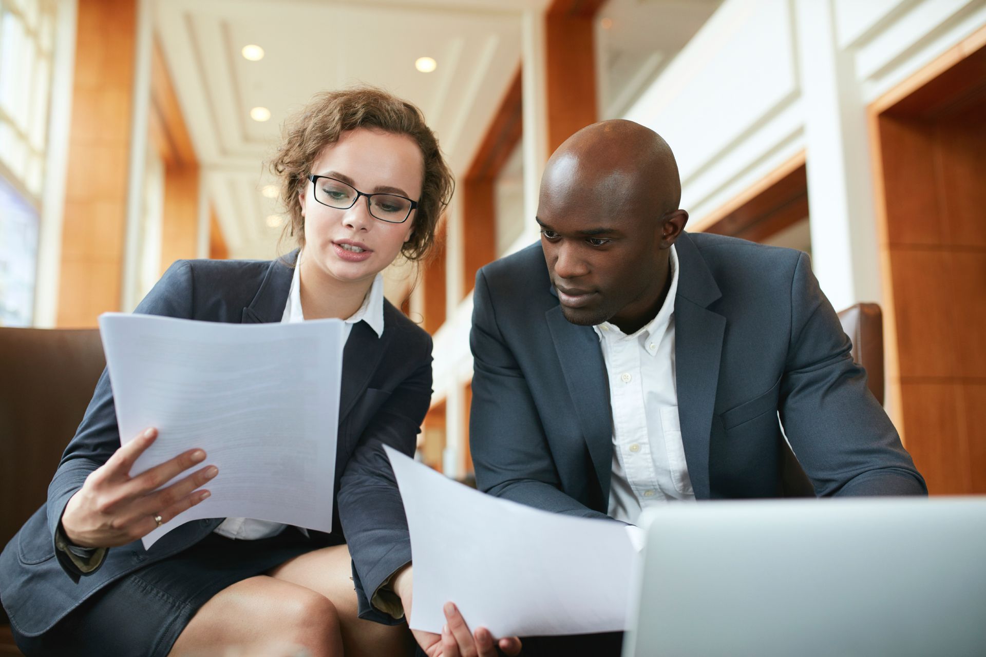 Two businesspeople in suits review documents, focused, near a laptop.