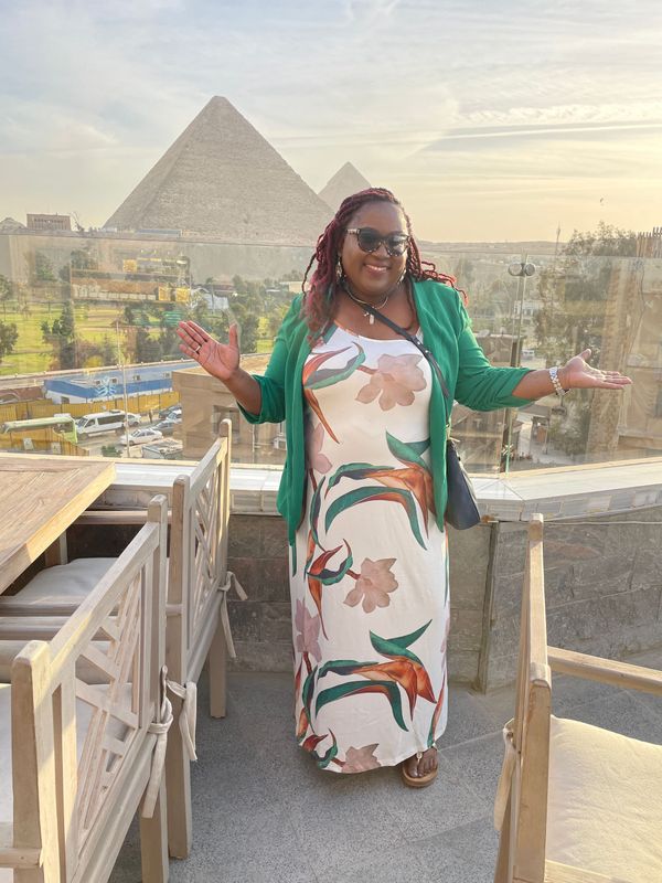 A woman in a white dress and green jacket is standing in front of the pyramids.