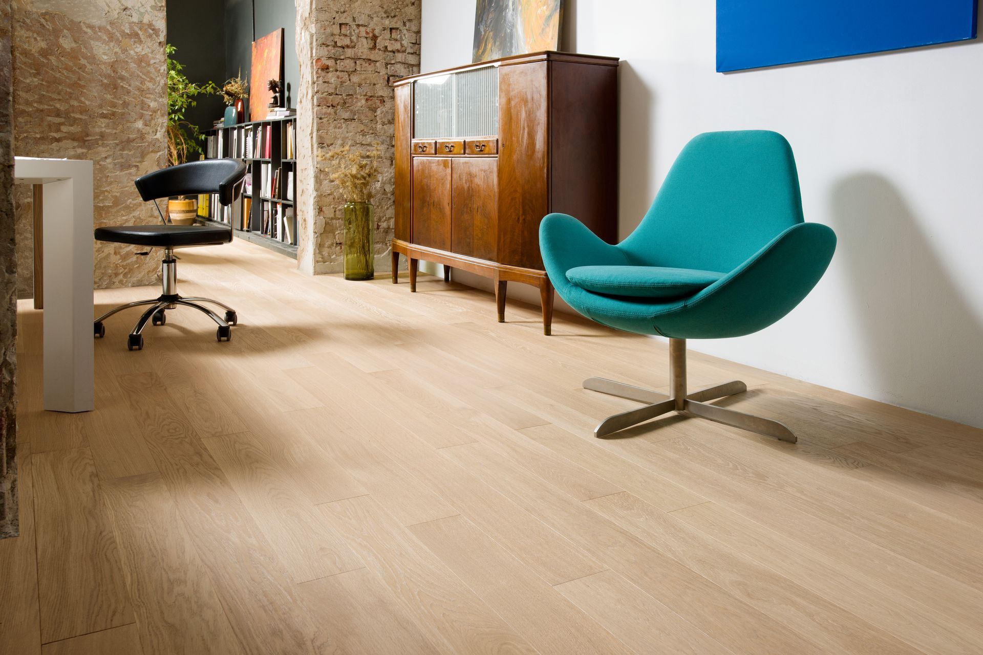 Wooden-floored room with teal chair, vintage cabinet, desk, bookshelf, and stone wall.
