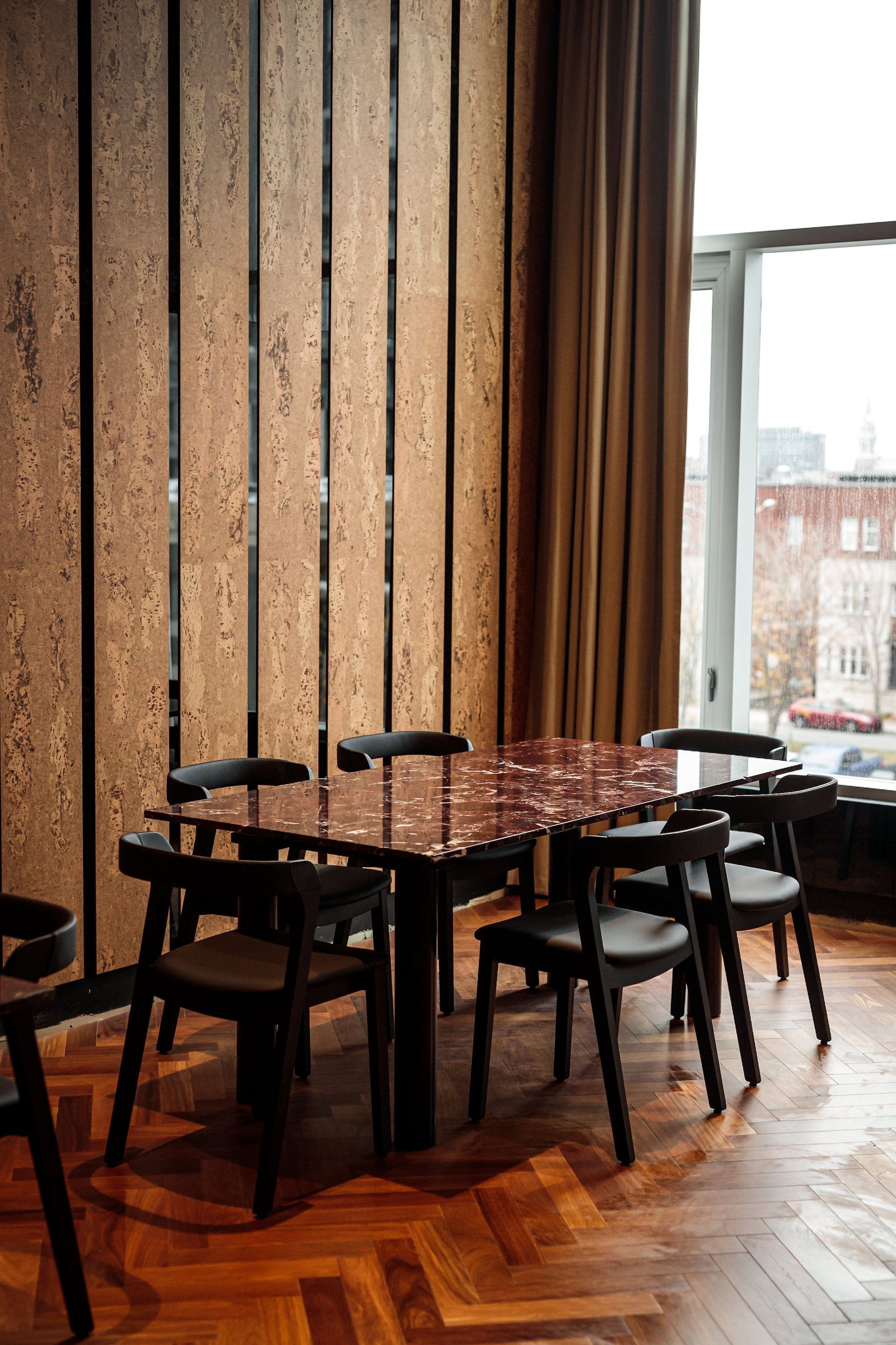 Dining table with six chairs, against a cork wall, by a large window with brown curtains.