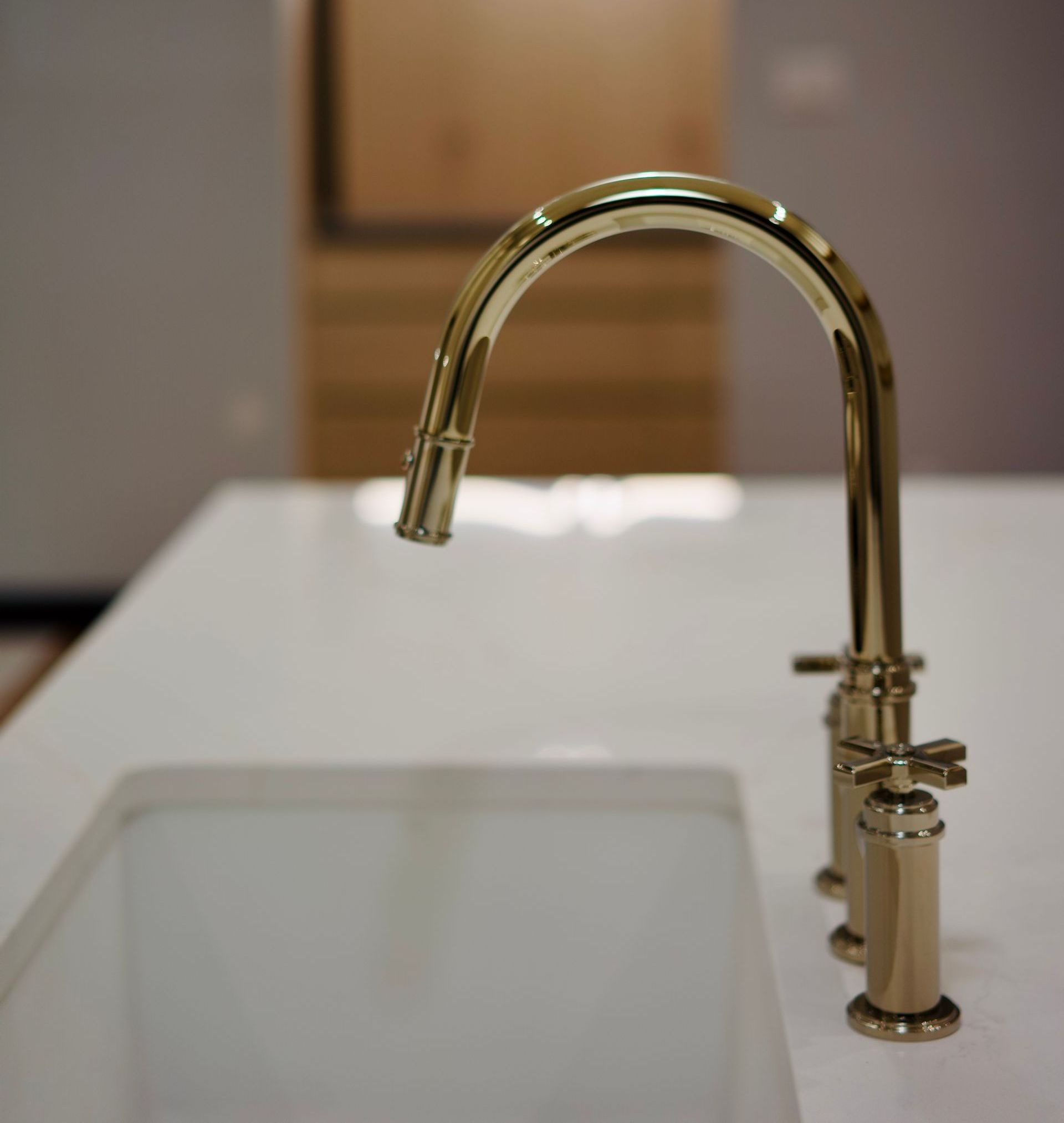 Robinet de cuisine doré au-dessus d'un évier blanc, posé sur un plan de travail blanc. L'arrière-plan est flou, laissant apparaître un mur et un meuble clairs.