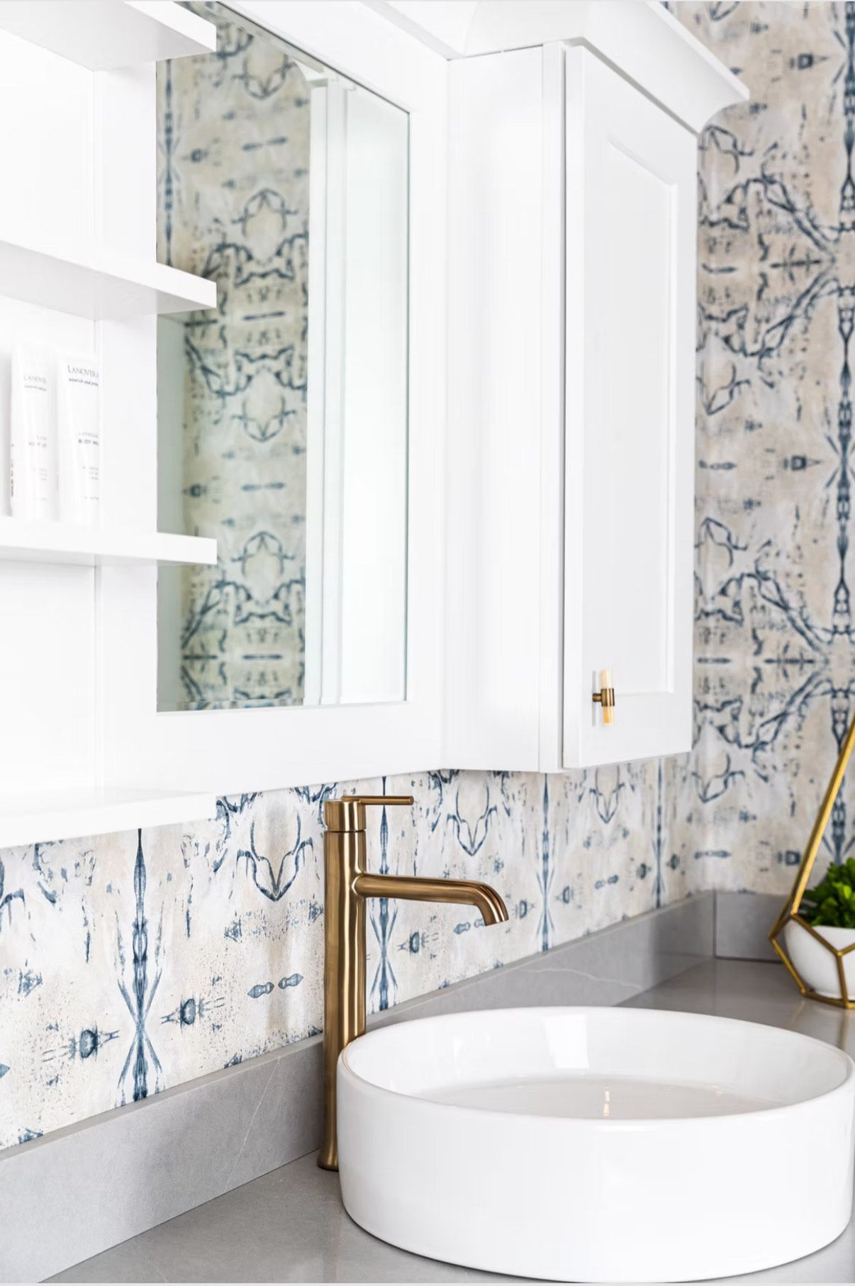 White bathroom vanity with gold faucet, round sink, and patterned wallpaper.