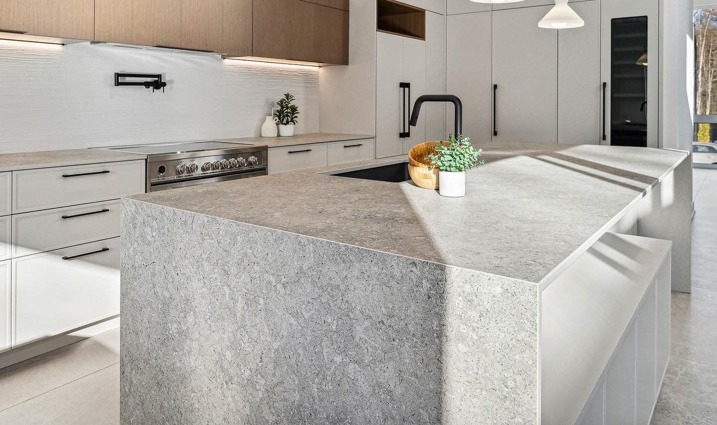 Cuisine moderne avec îlot en pierre grise, armoires blanches et touches de bois. Comprend une cuisinière, un robinet et une petite plante.