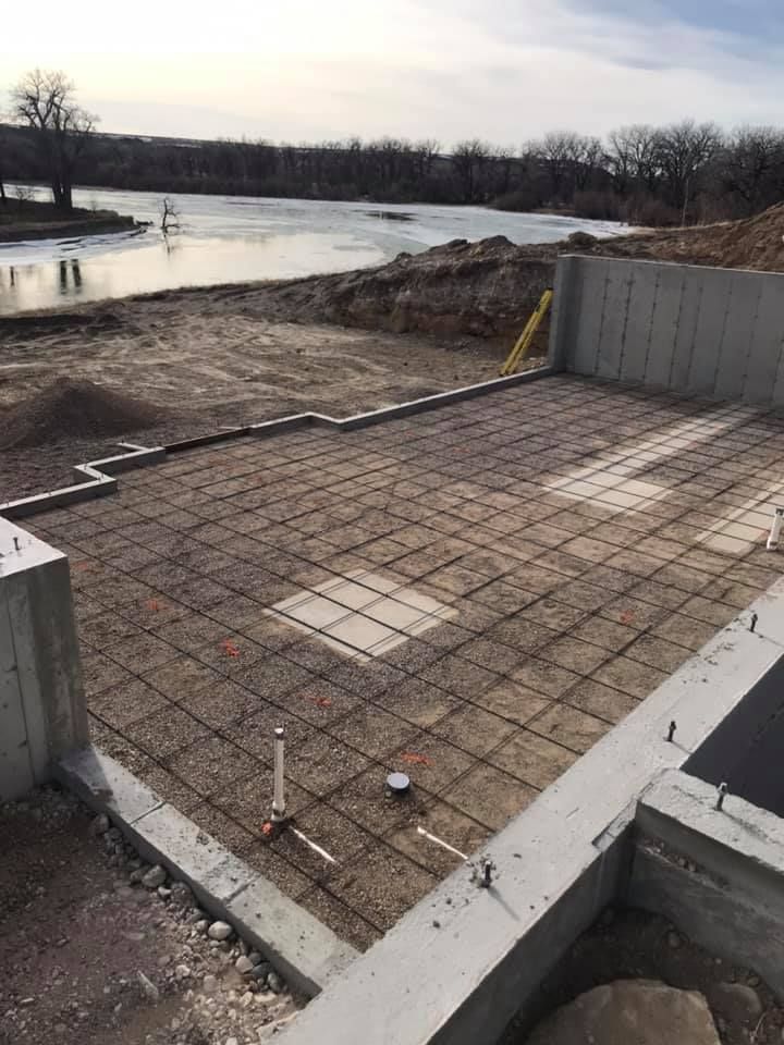 Construction site with concrete foundation, rebar grid, and nearby water.
